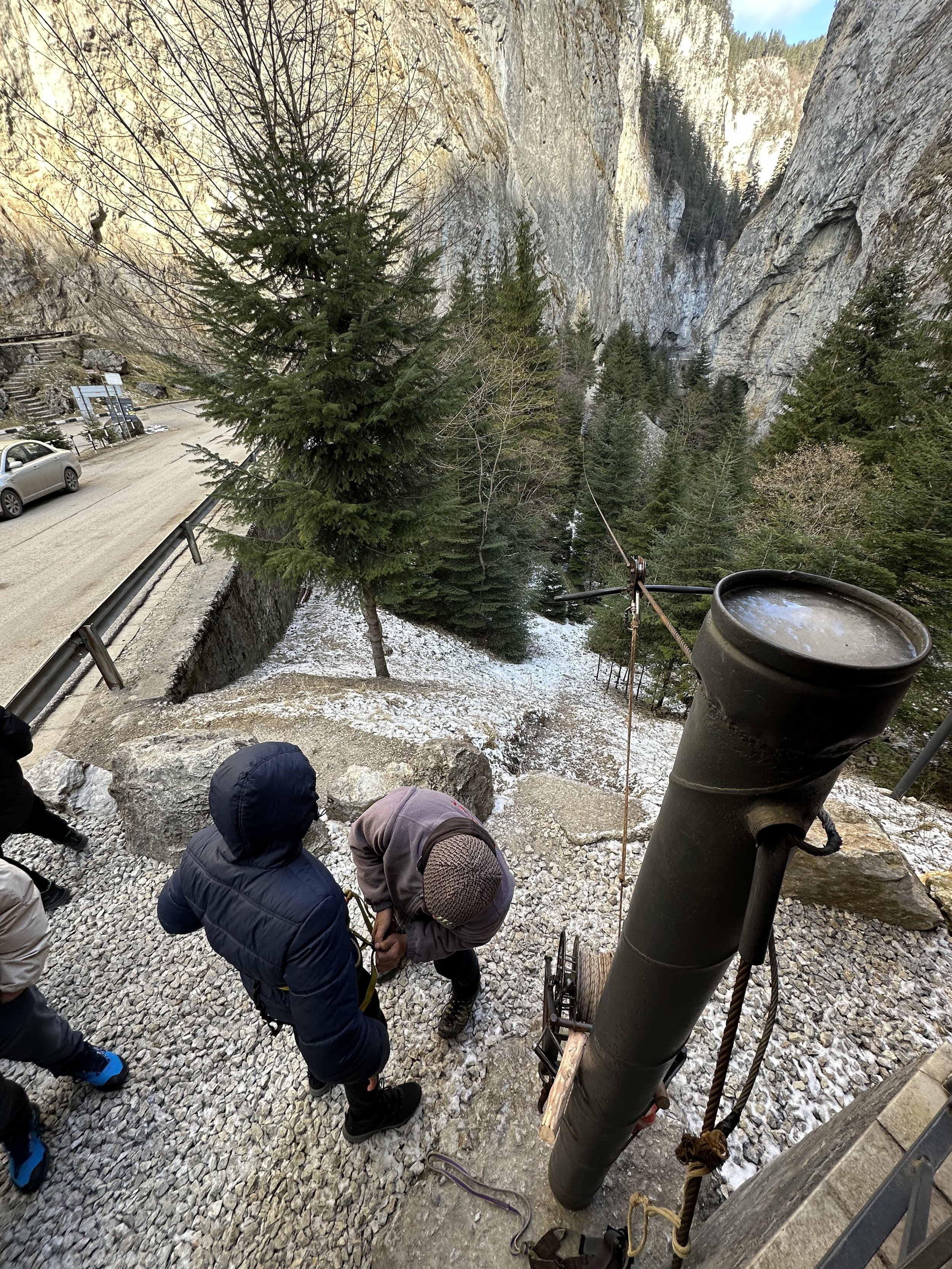 People near a canyon wearing winter clothes, with a safety harness being adjusted, surrounded by trees and rocky cliffs.