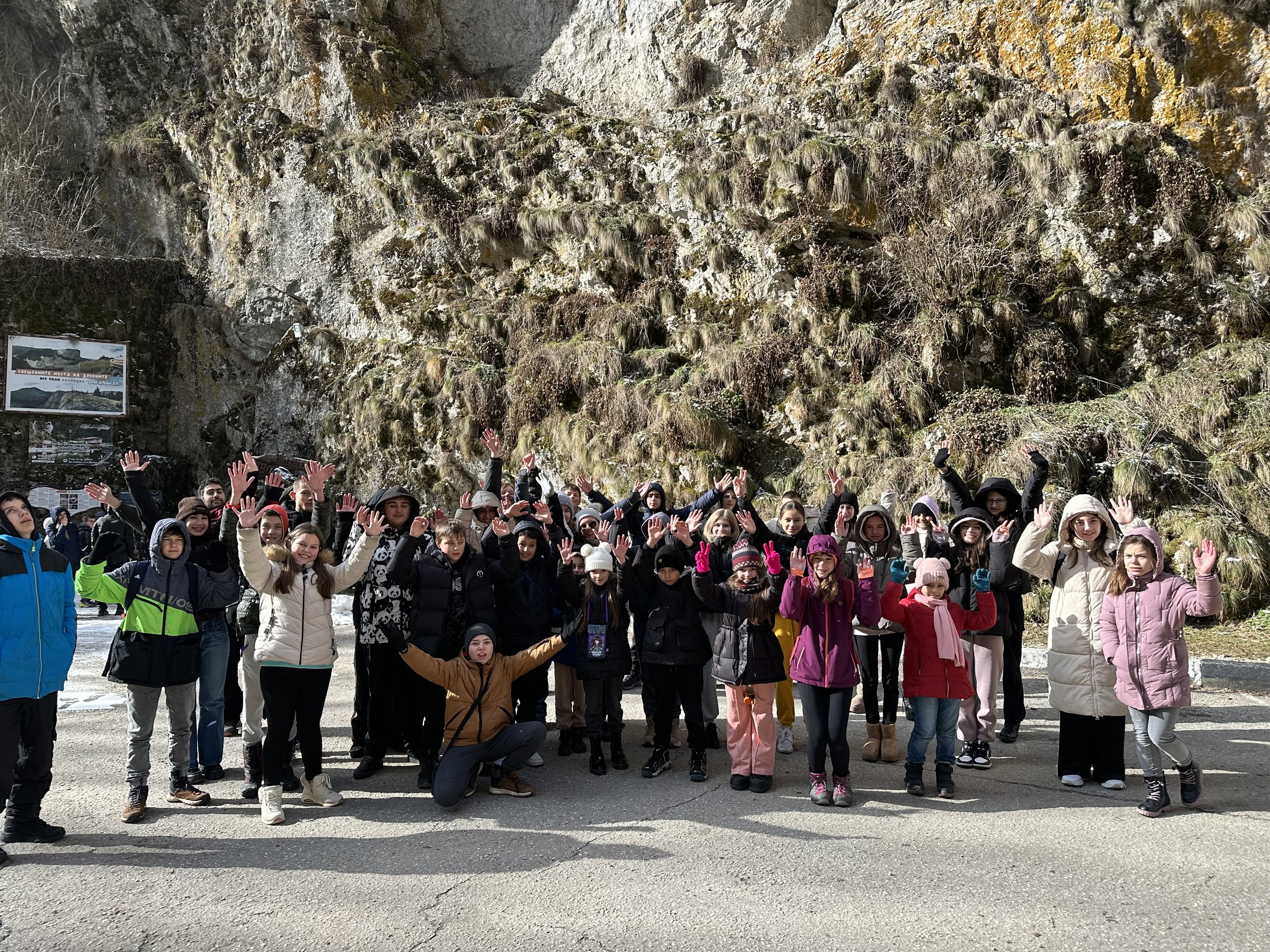 A group of people wearing winter clothing, standing outdoors in front of a rocky hillside. They have raised their arms, waving at the camera, and appear to be enjoying a sunny day.