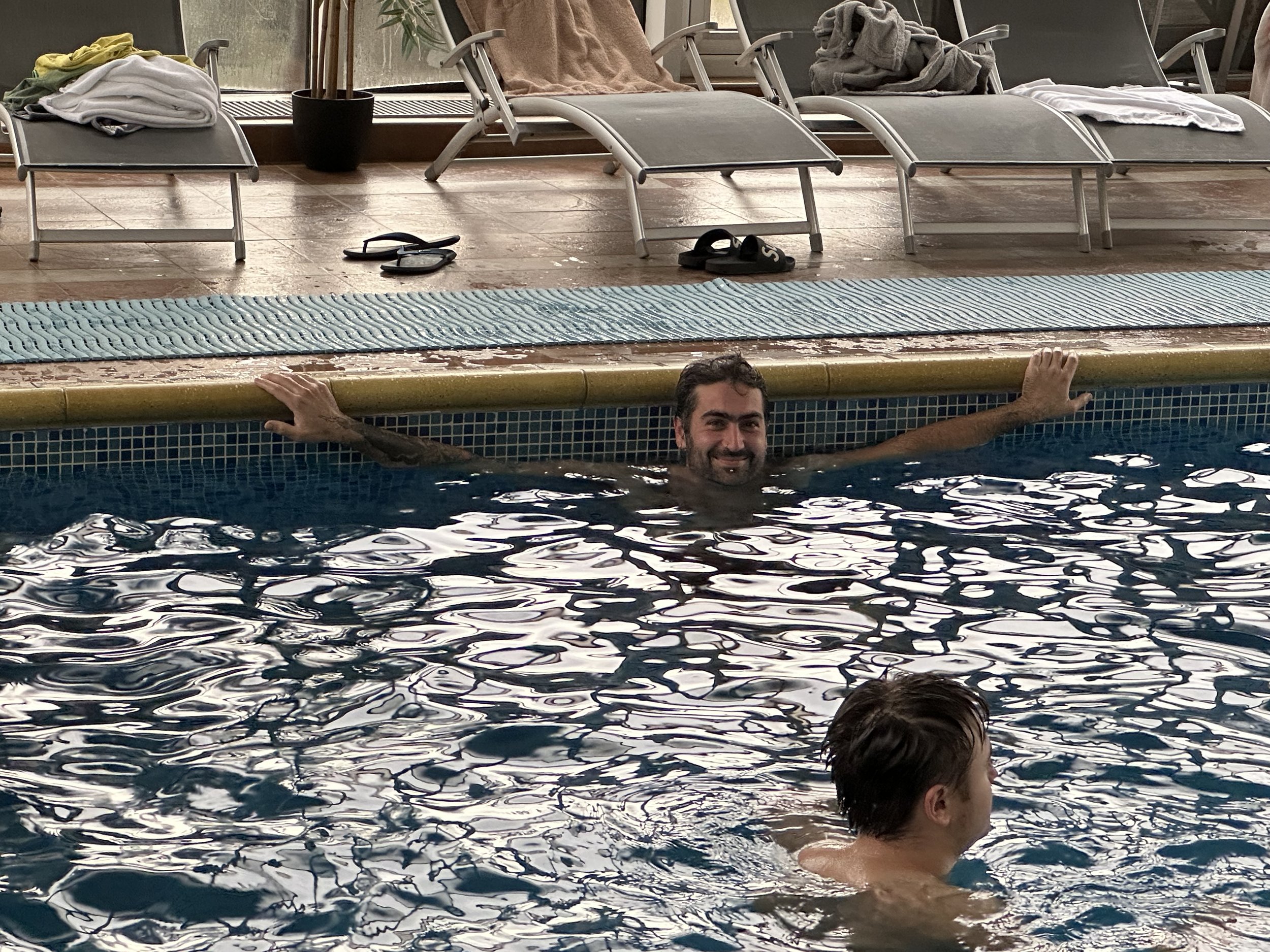 People swimming in an indoor swimming pool with lounge chairs and towels in the background.