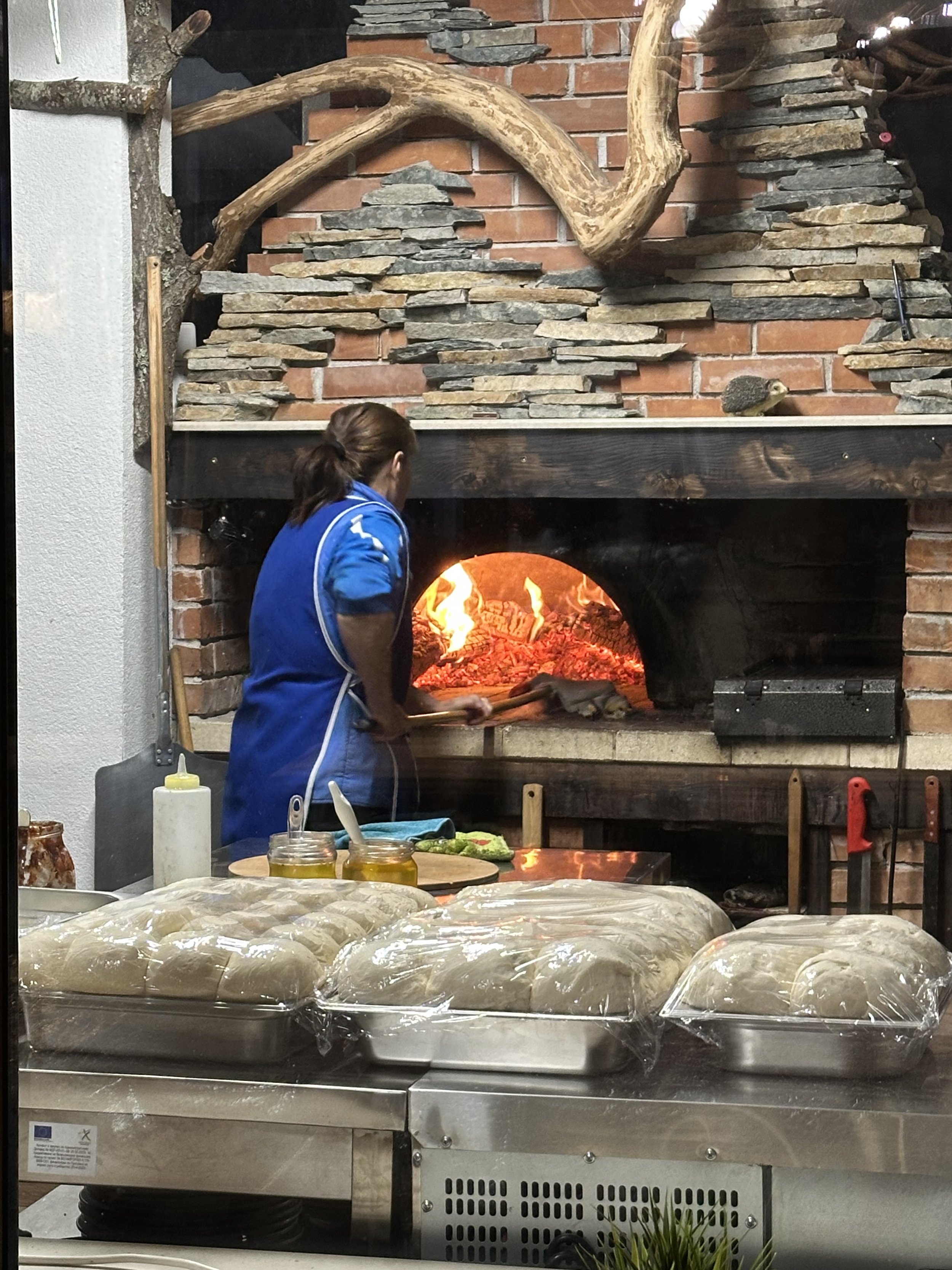 Person in blue attire baking pizza in a wood-fired oven with dough trays in the foreground.