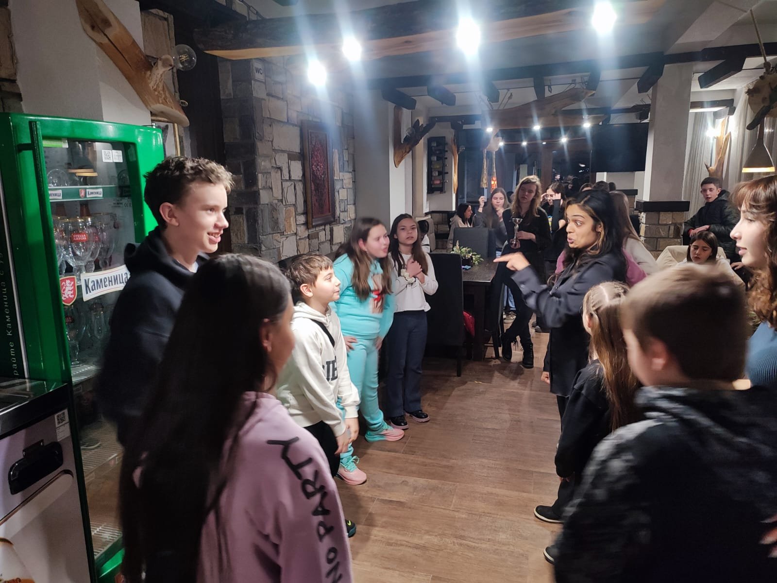 A group of children standing in a circle, guided by an adult woman, inside a cozy room with wooden floors and stone walls. A refrigerator with glass doors is visible on the left.