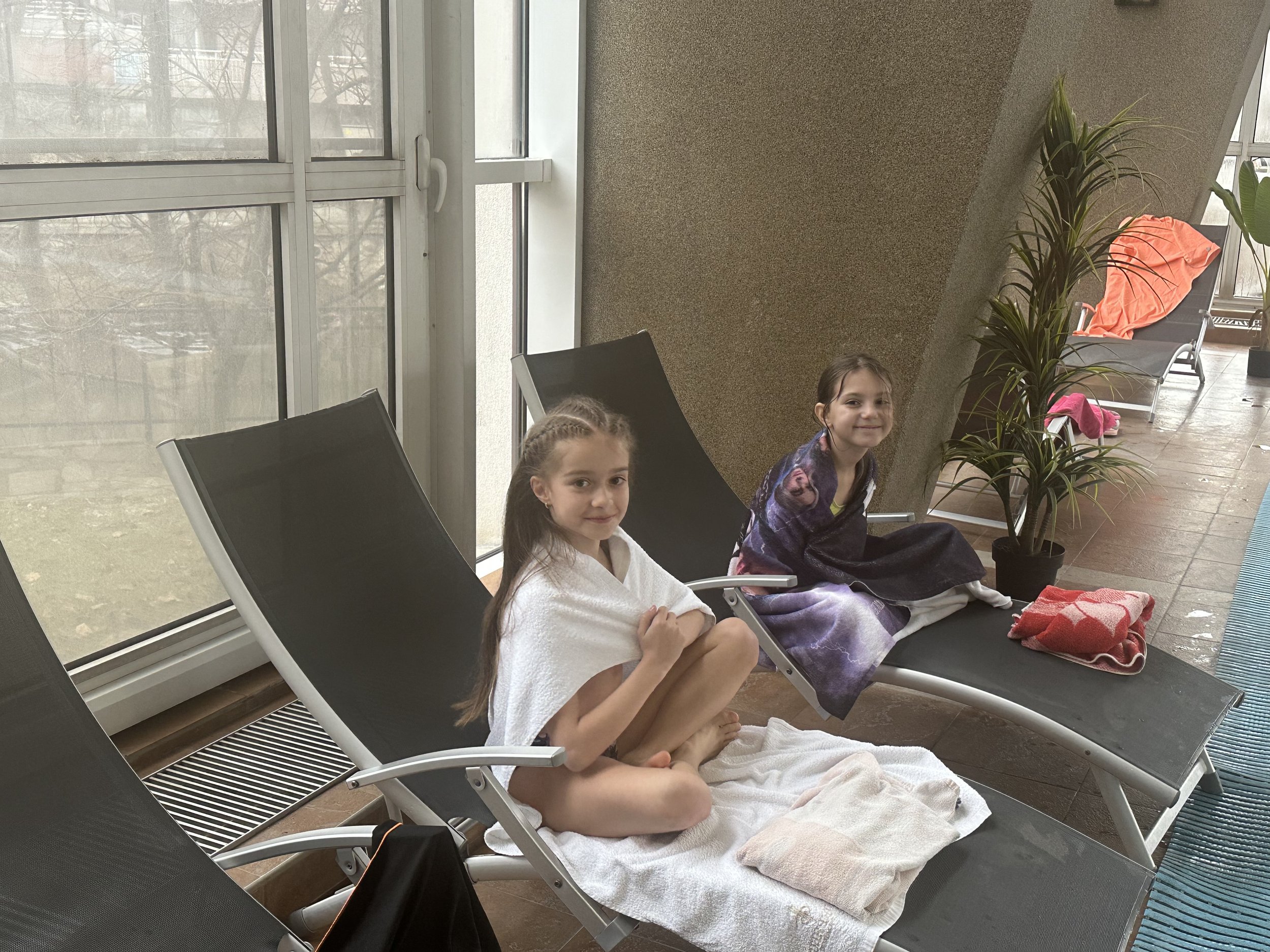 Two young girls sitting on poolside lounge chairs wrapped in towels indoors with large windows in the background.