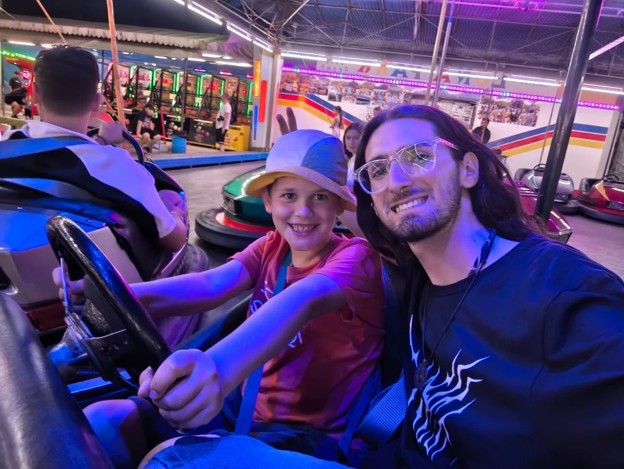A smiling boy and man taking a selfie while riding bumper cars at an amusement park. The boy is wearing a bucket hat and a red shirt, and the man has long hair, glasses, and a septum piercing. The background features colorful lights and bumper cars.