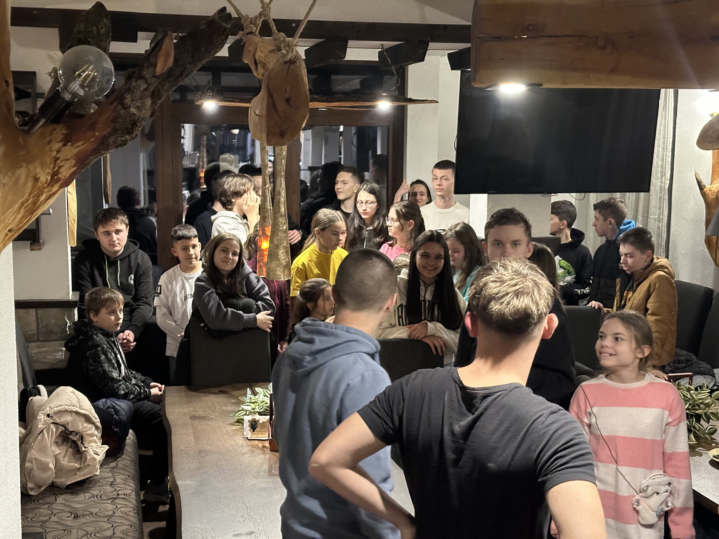 A group of people, mostly young, in a room with wooden beams and a mix of casual clothing. The room has modern lighting fixtures and some rustic wood decor. They appear to be gathered informally, some sitting and others standing, engaging in conversa