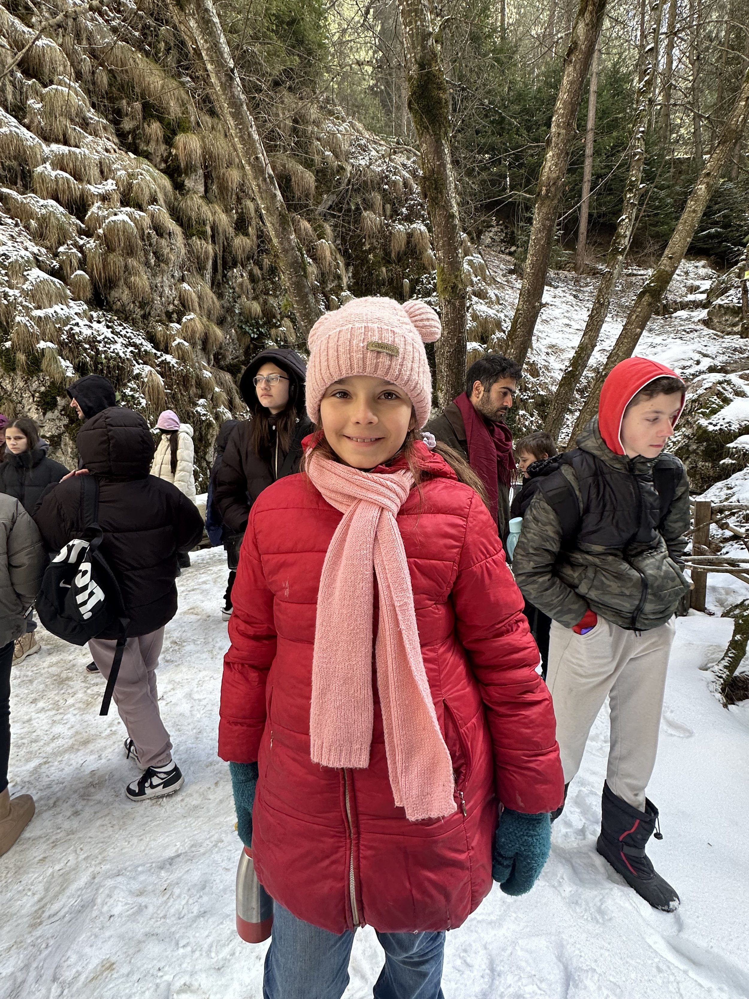 A young girl in a red coat and pink hat smiles in a snowy forest, surrounded by other people dressed warmly.