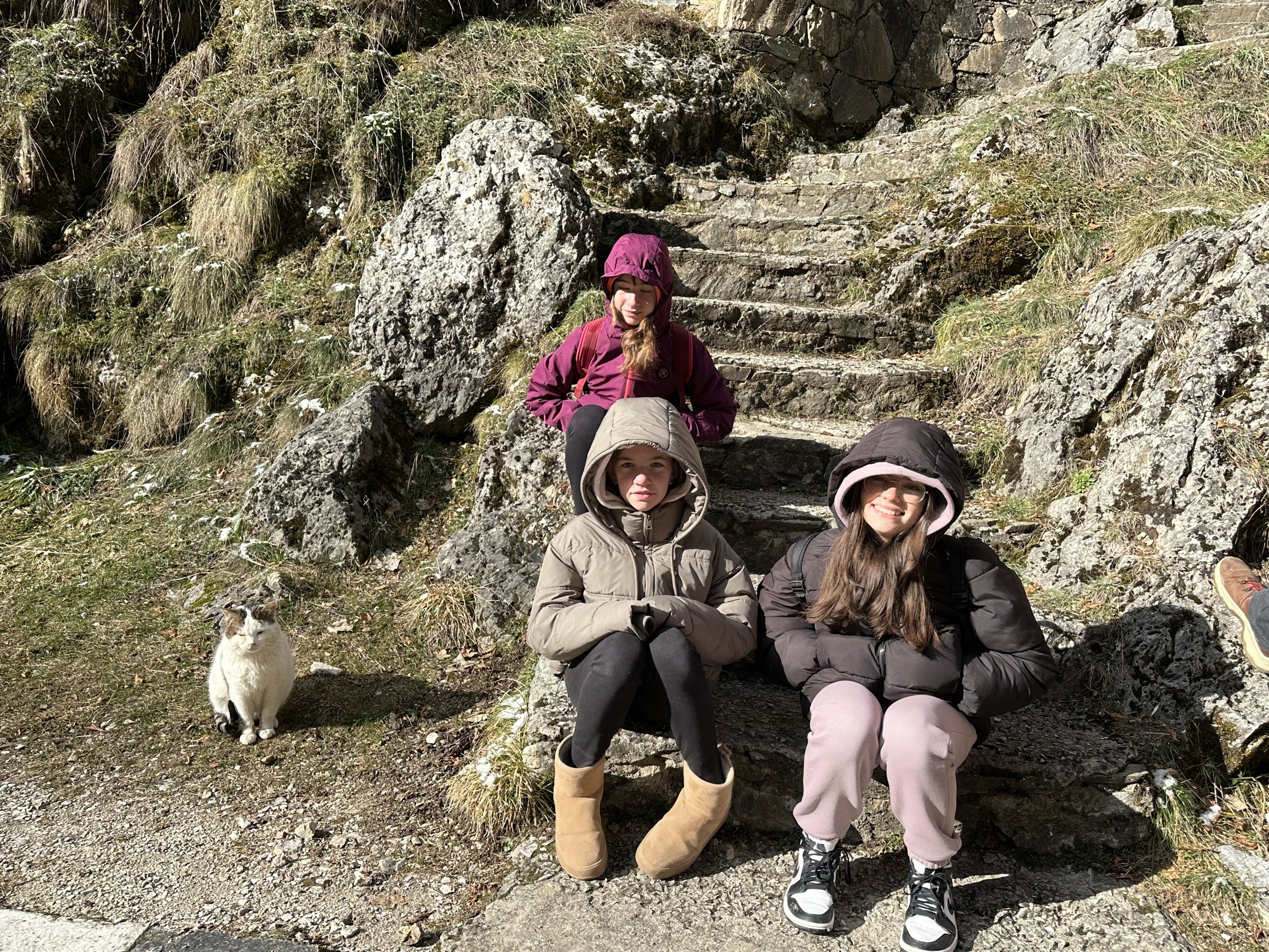 Three people wearing winter jackets sitting on stone steps in a rocky, grassy outdoor setting, with a cat sitting nearby.