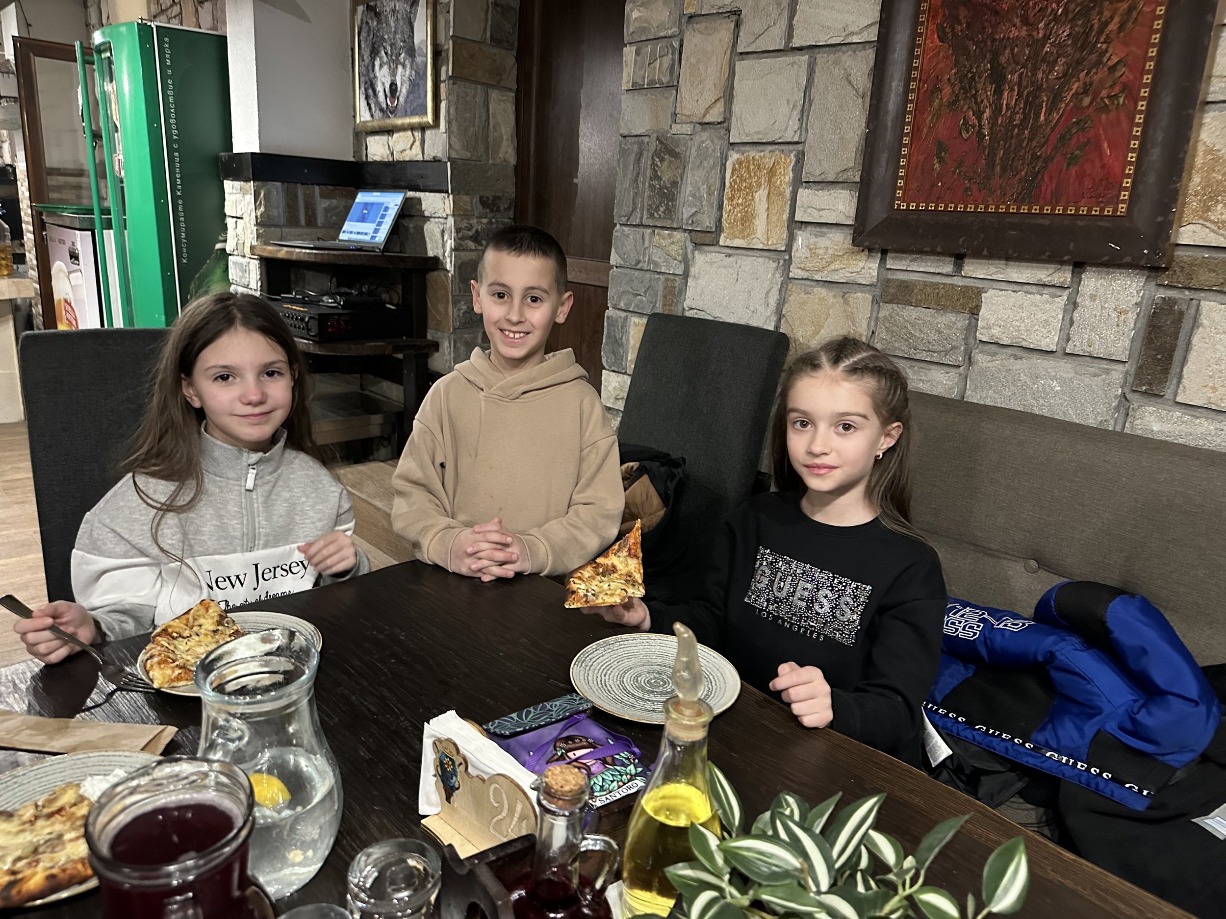 Three children sitting at a table in a restaurant, each with a slice of pizza on their plate. Two girls and one boy are smiling. The table has a jug of water and some condiments. Stone walls and artwork are visible in the background.