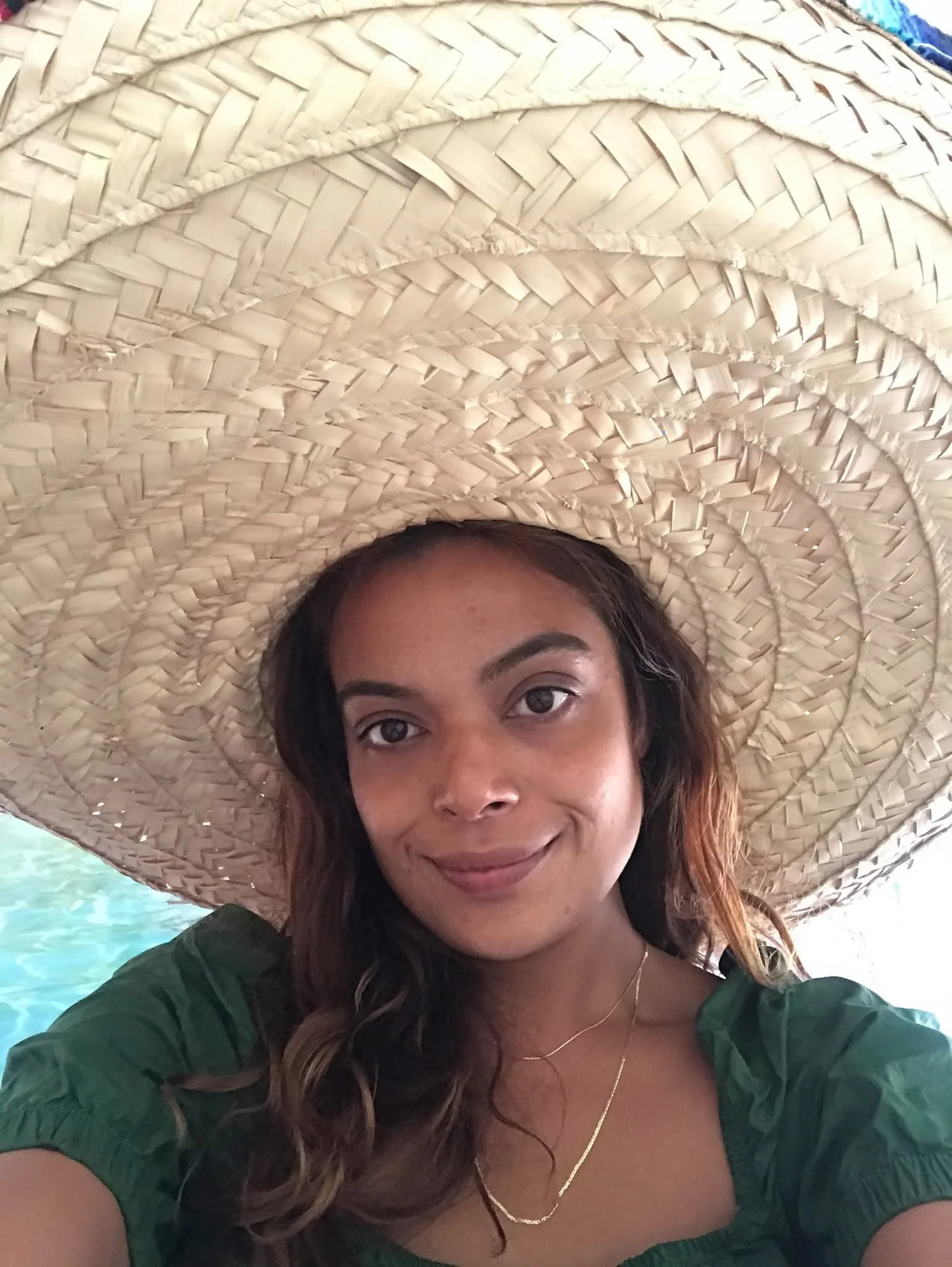 A woman with curly brown hair and medium skin tone wearing a large sun hat, green top, and a delicate gold necklace, smiling outdoors near water.