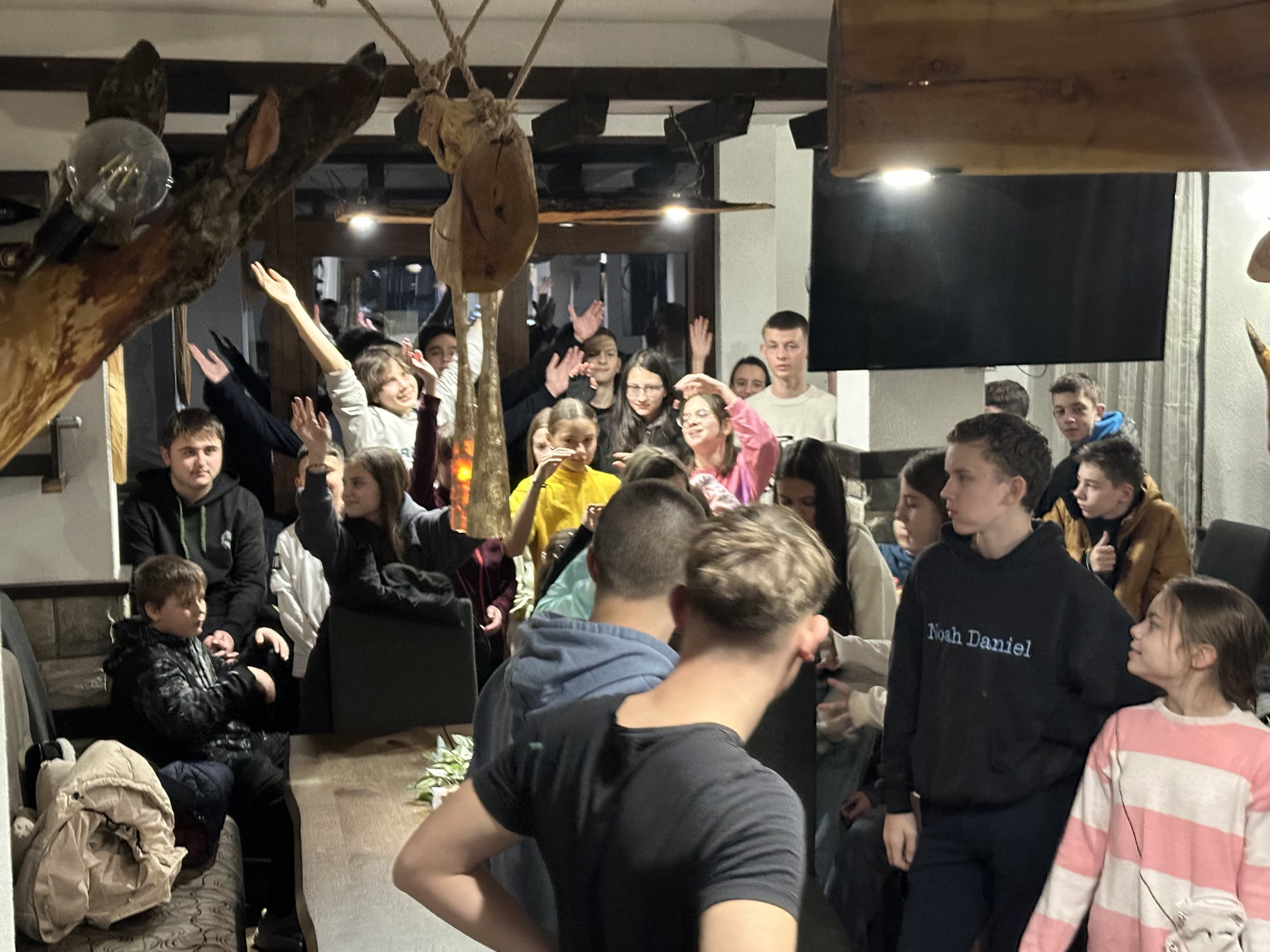 Group of people gathered in a room, some raising their hands. Young men and women are sitting and standing around a table, engaging with each other. The room has wooden decor and lighting fixtures.