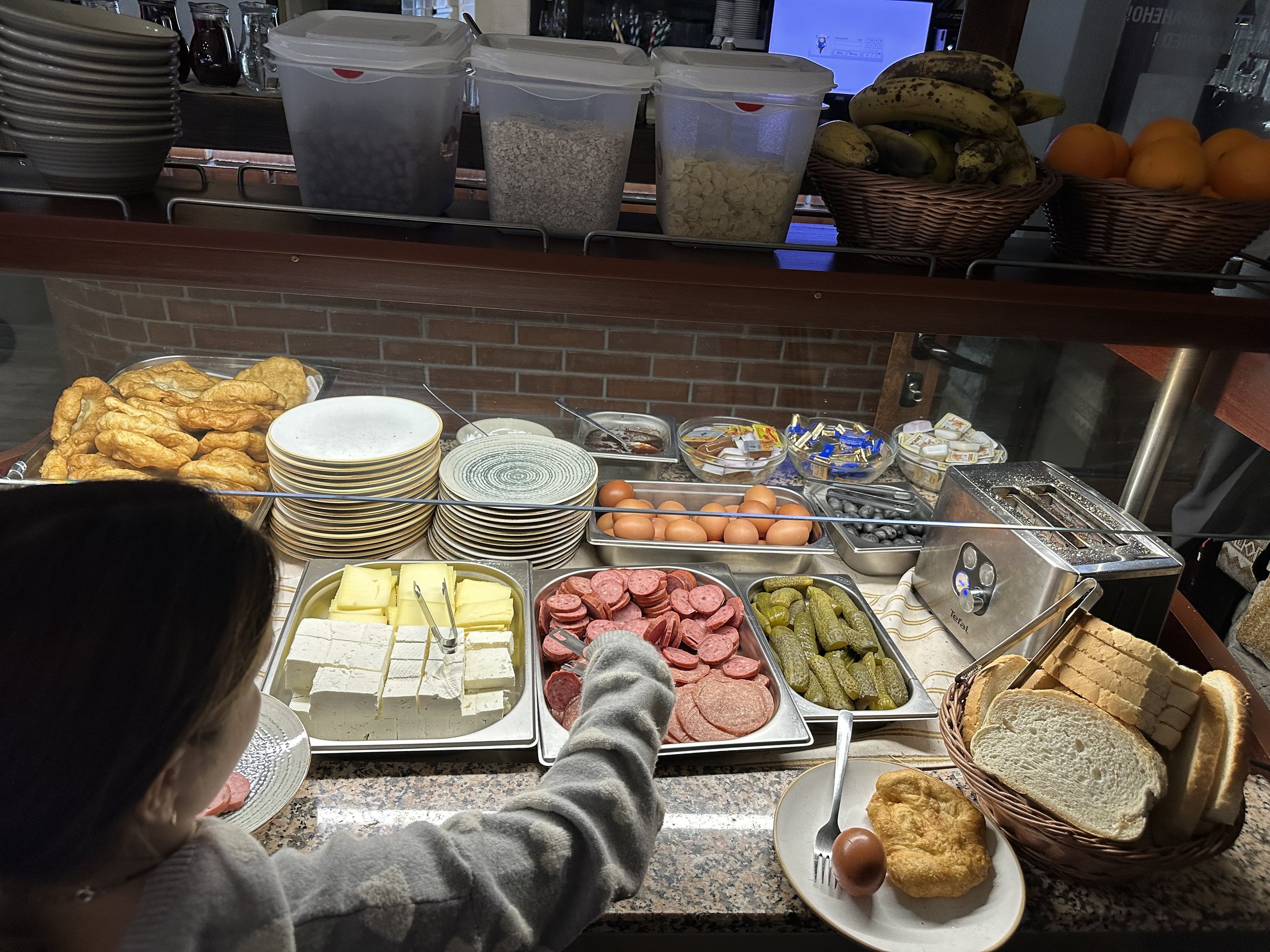 Buffet breakfast with pastries, cheese, cold cuts, boiled eggs, pickles, bread, bananas, and oranges.