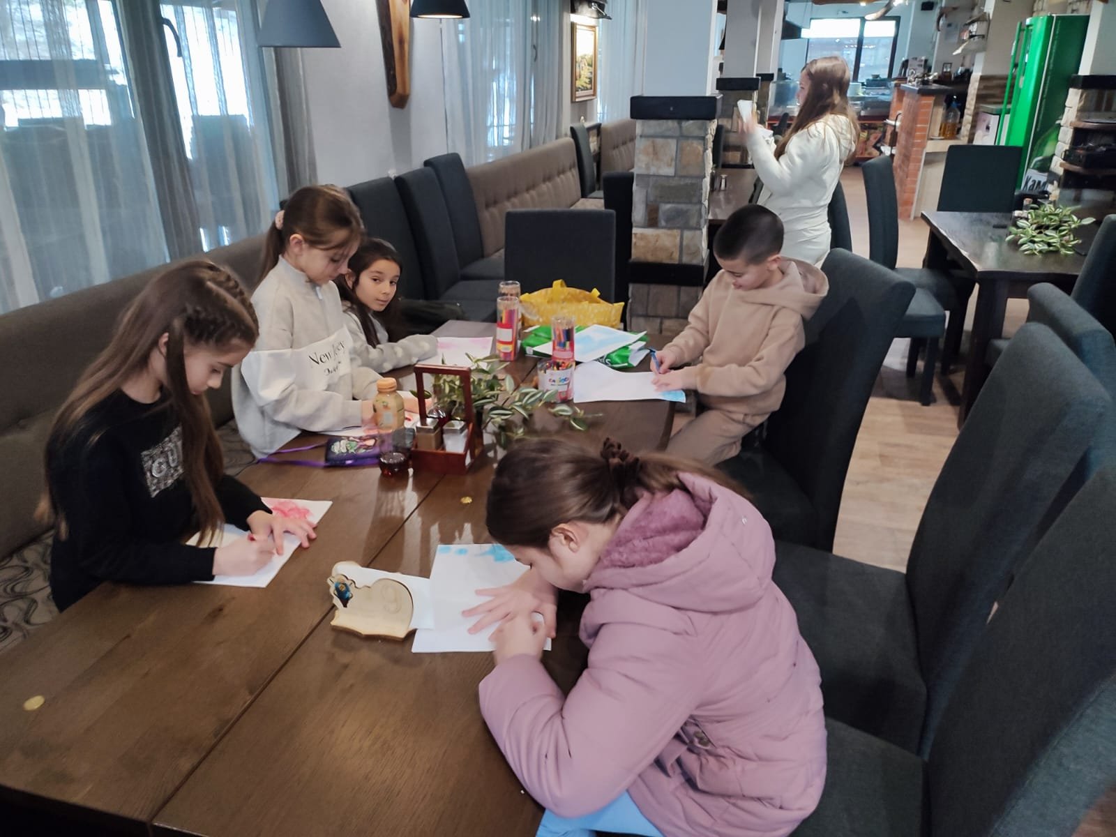 Children sitting at a long table in a restaurant, engaged in drawing and writing activities with papers, markers, and colored pencils. In the background, an adult is standing, looking at a phone.