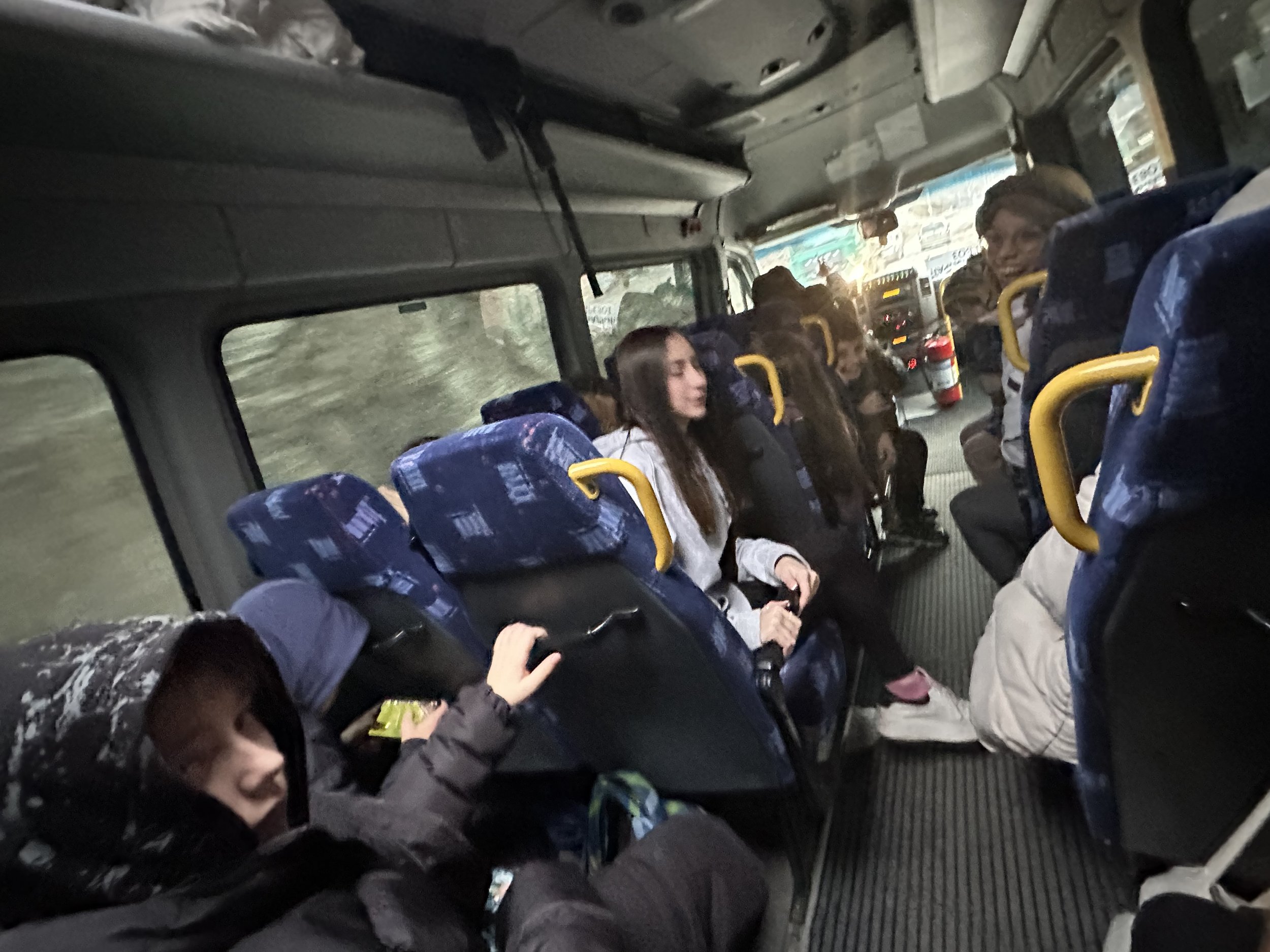 Passengers sitting inside a bus with blue seats, wearing winter clothing.