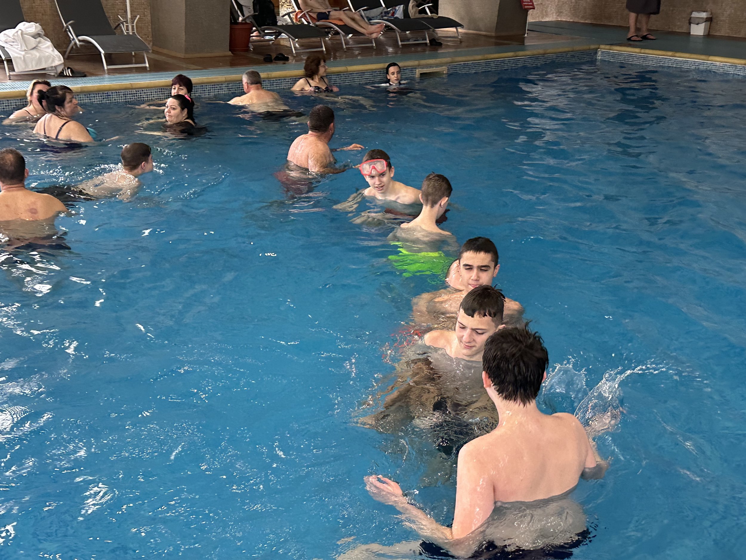 Indoor swimming pool with several people swimming and socializing, including adults and children, some wearing goggles. Poolside chairs visible in the background.