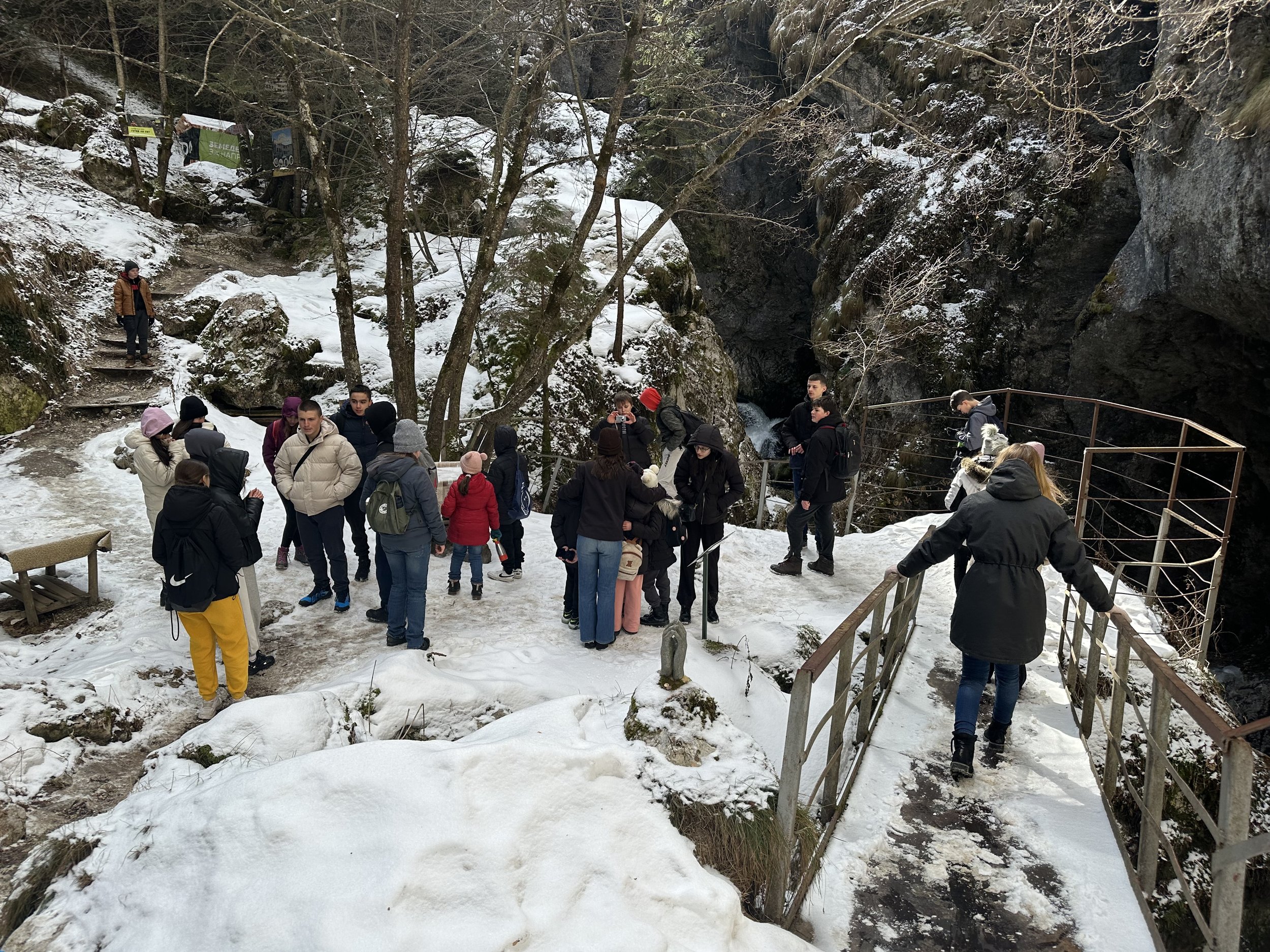 A group of people in winter clothing standing on a snowy walkway in a forested area with rocky terrain. Some individuals are gathered near a small bridge while others stand on a path with snow-covered trees and rocks in the background.