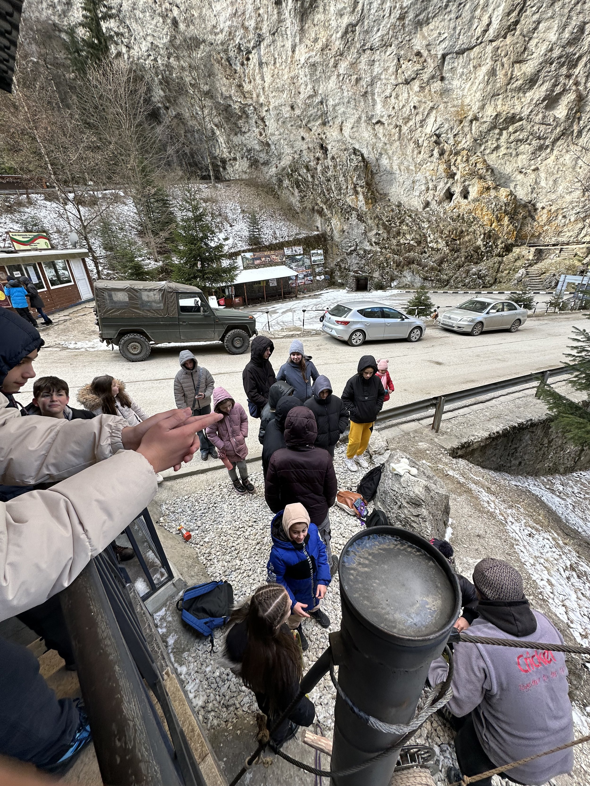 Group of people standing on a rocky path near parked vehicles, with a steep cliff face and snow-dusted trees in the background.
