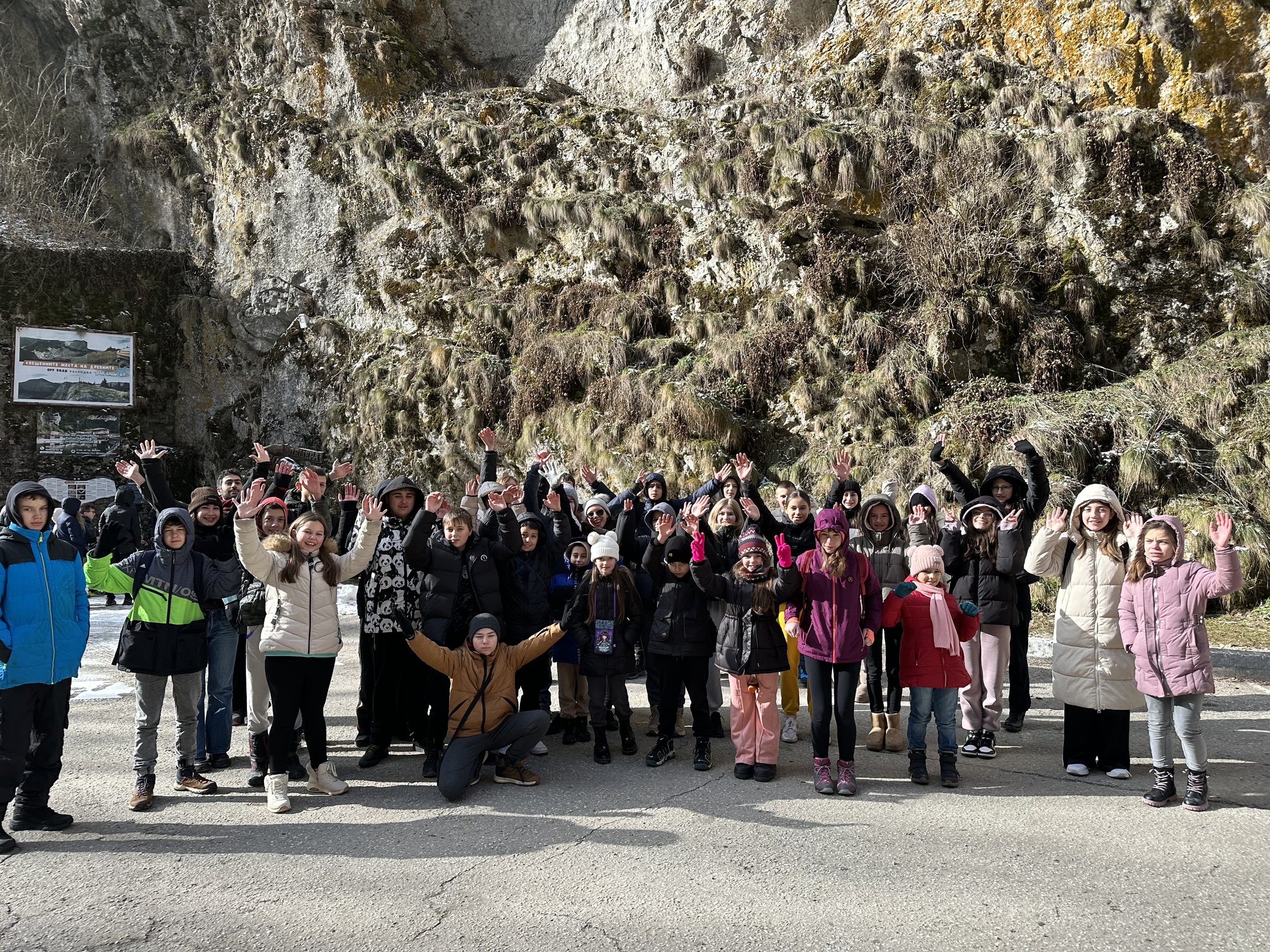 A group of people wearing winter clothing, standing outdoors with their hands raised, in front of a rocky, moss-covered cliff.