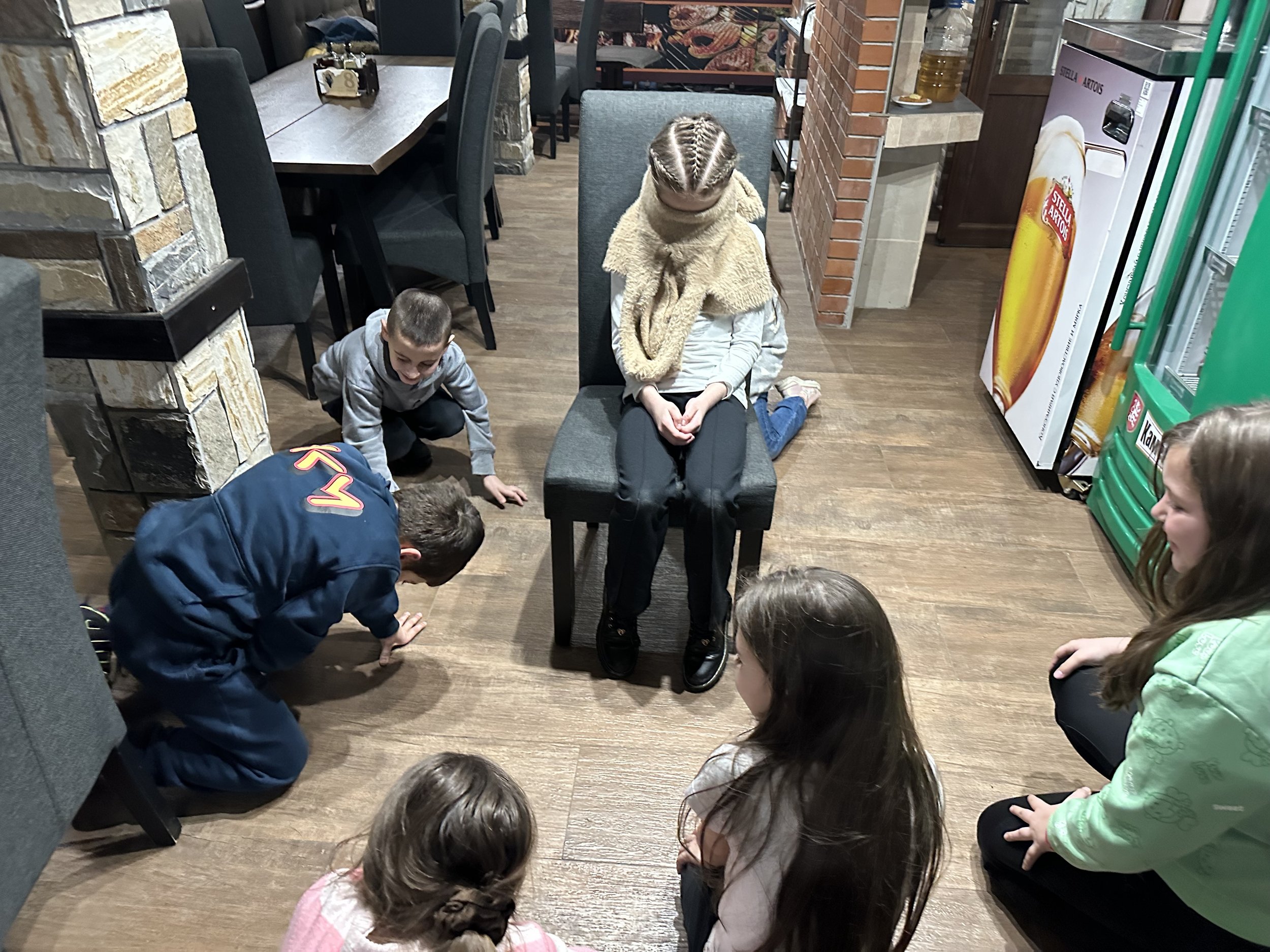 Children gathered around a girl sitting on a chair with a scarf covering her face, in a room with stone and brick walls, tables, and a refrigerator displaying a beer advertisement.