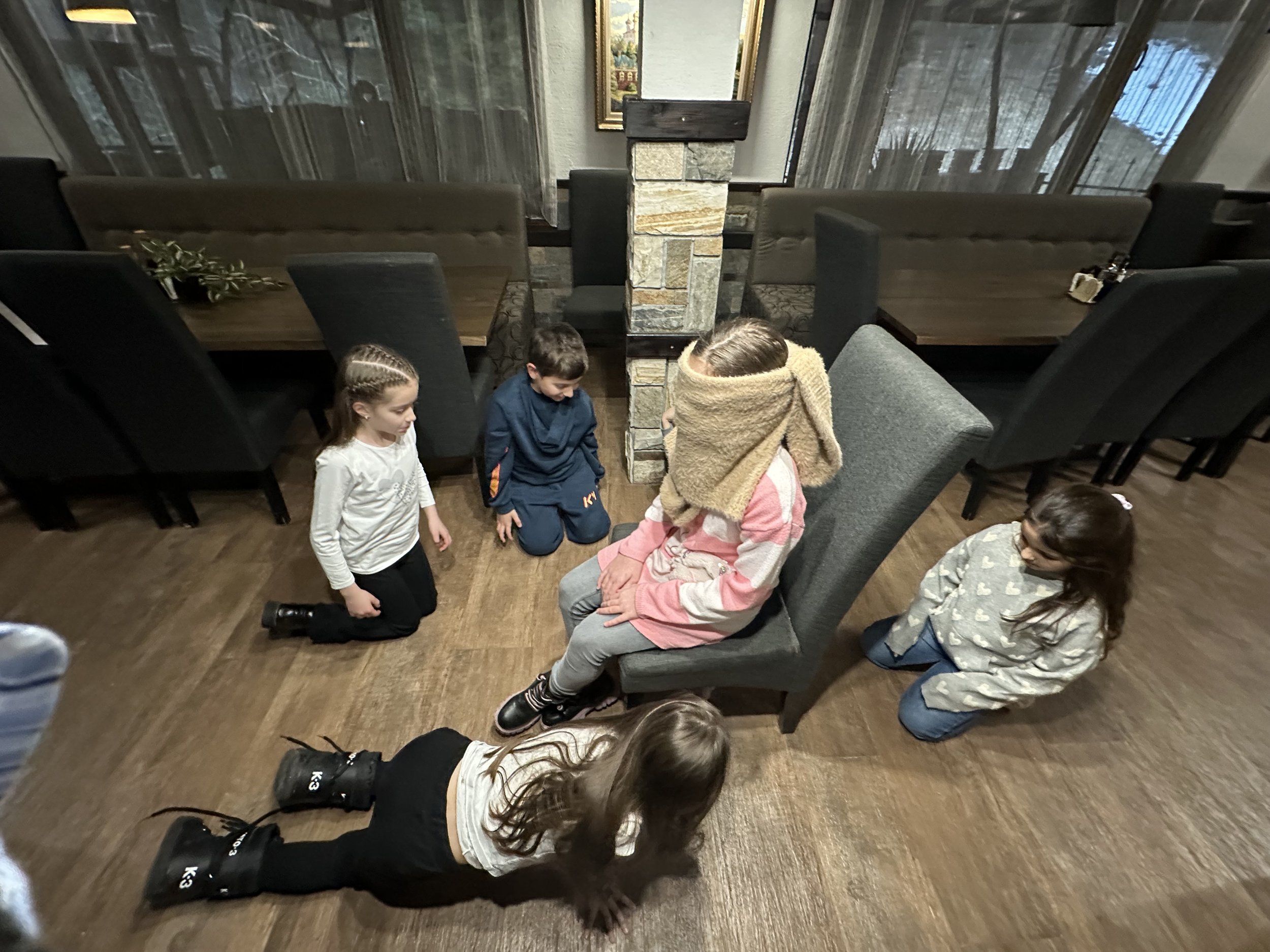 Children playing a guessing game indoors with one child blindfolded on a chair and others kneeling nearby.