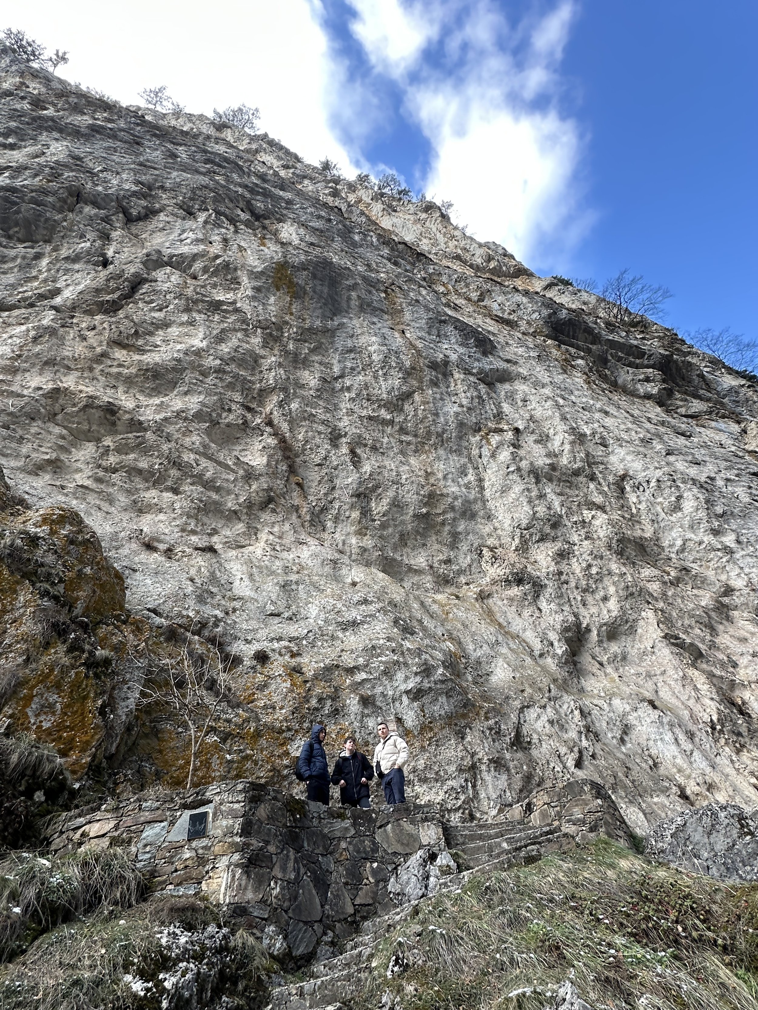 Three people standing on stone steps next to a large rock face under a blue sky with clouds.