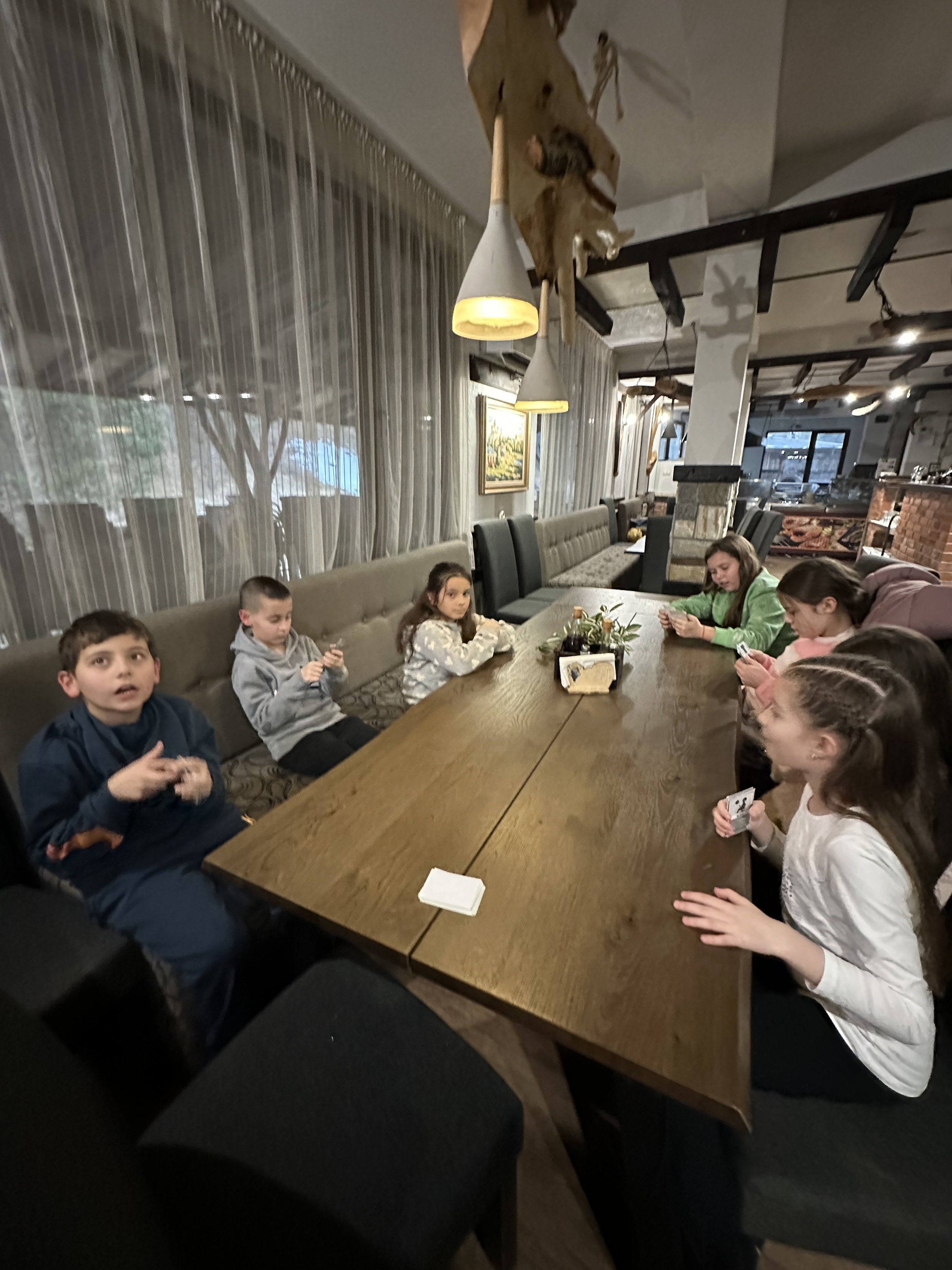 Children sitting around a wooden table in a restaurant setting, playing a card game. The room has modern pendant lights, decorative curtains, and upholstered benches.