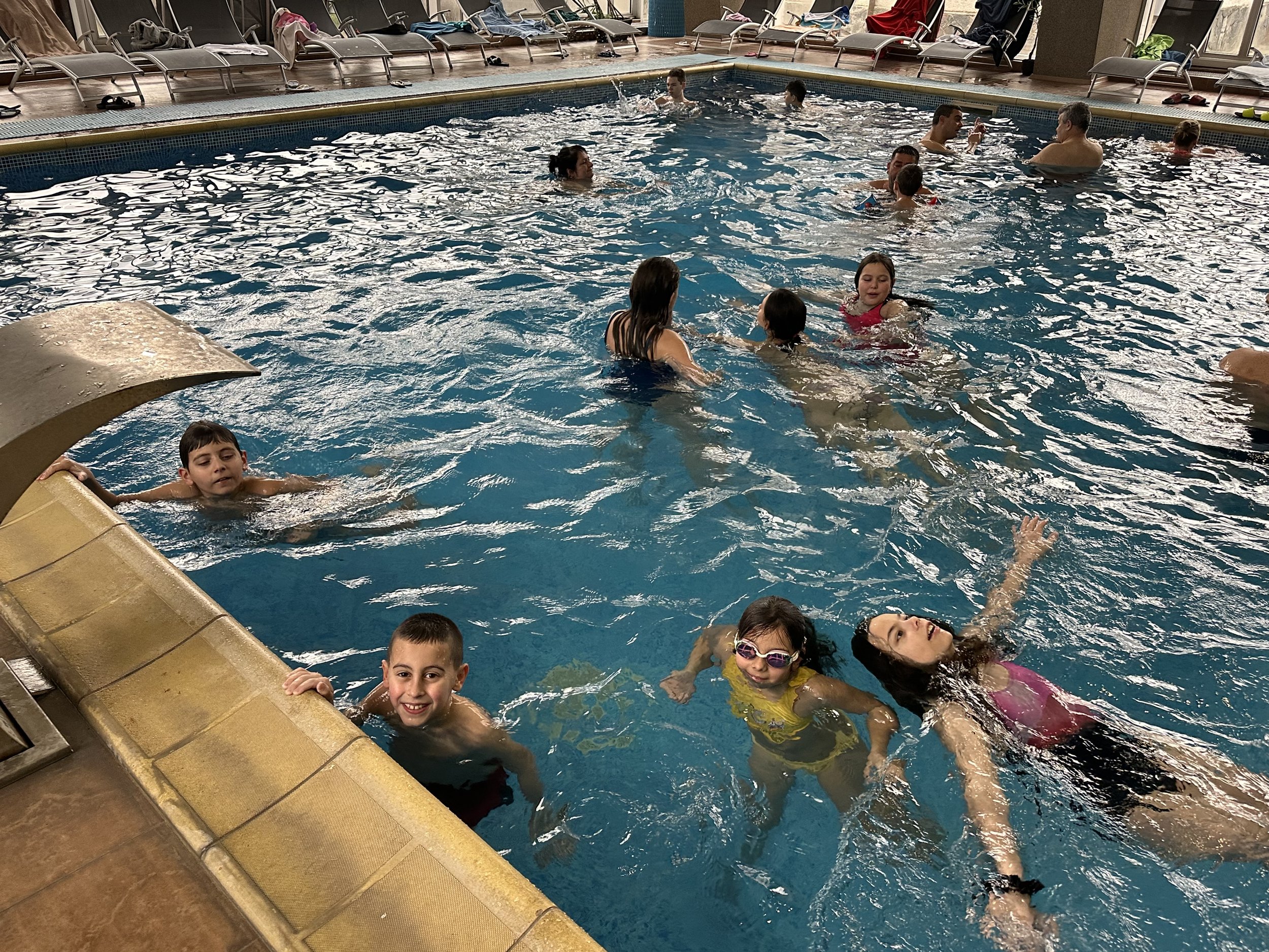 Indoor swimming pool with several people enjoying the water, including children and adults, with a row of lounge chairs in the background.