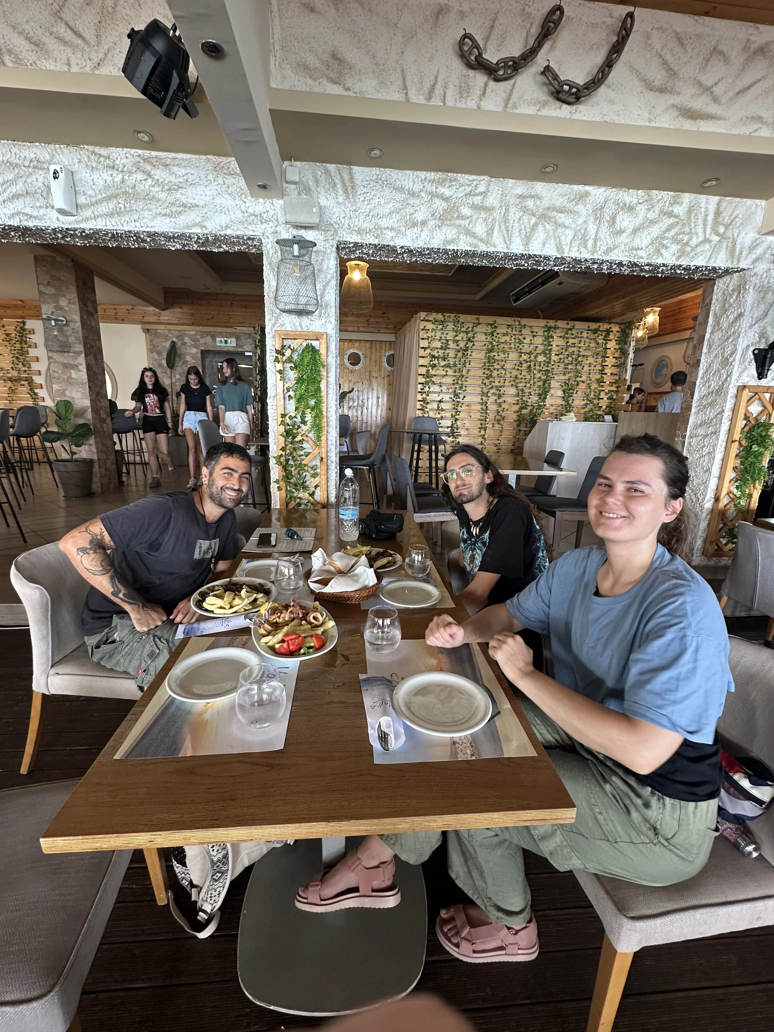 Camp Evolve International instructors enjoying lunch together at a restaurant in Kavala, Greece, sitting at a table with food and drinks and smiling during a relaxed break.