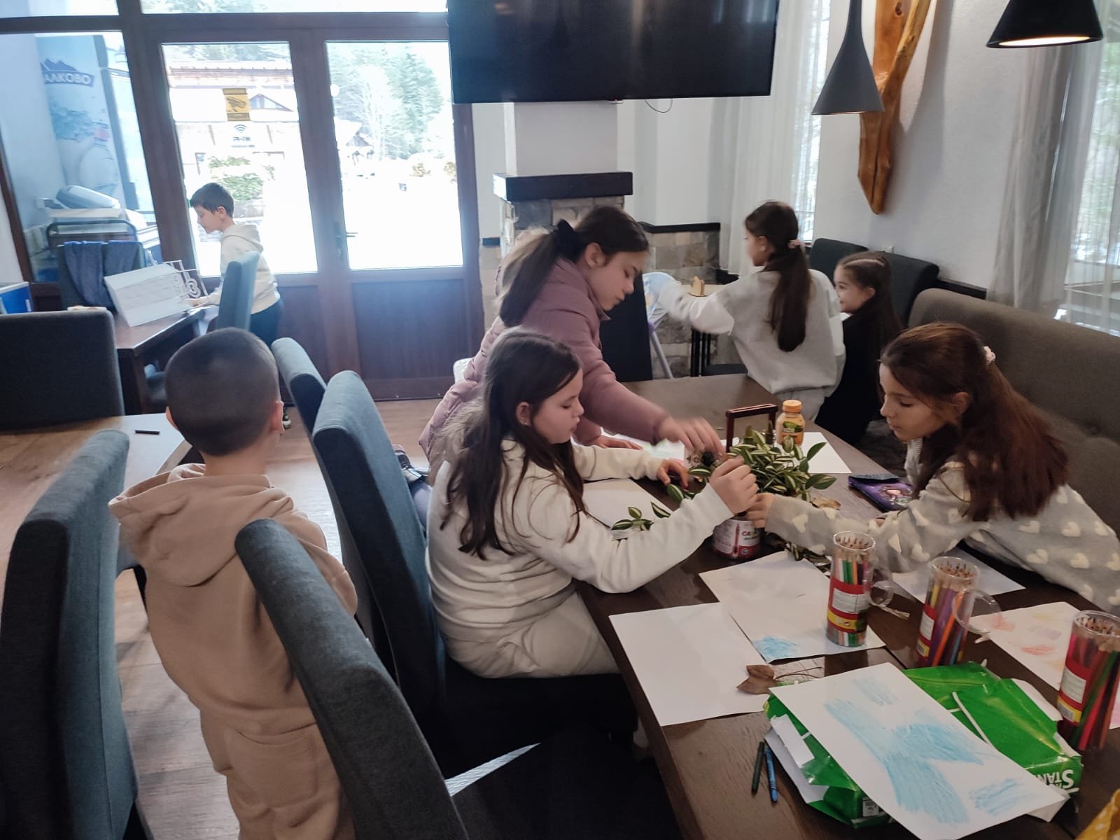 Children engaging in a creative activity at a table with drawings, colored pencils, and plant cuttings in a room with large windows.