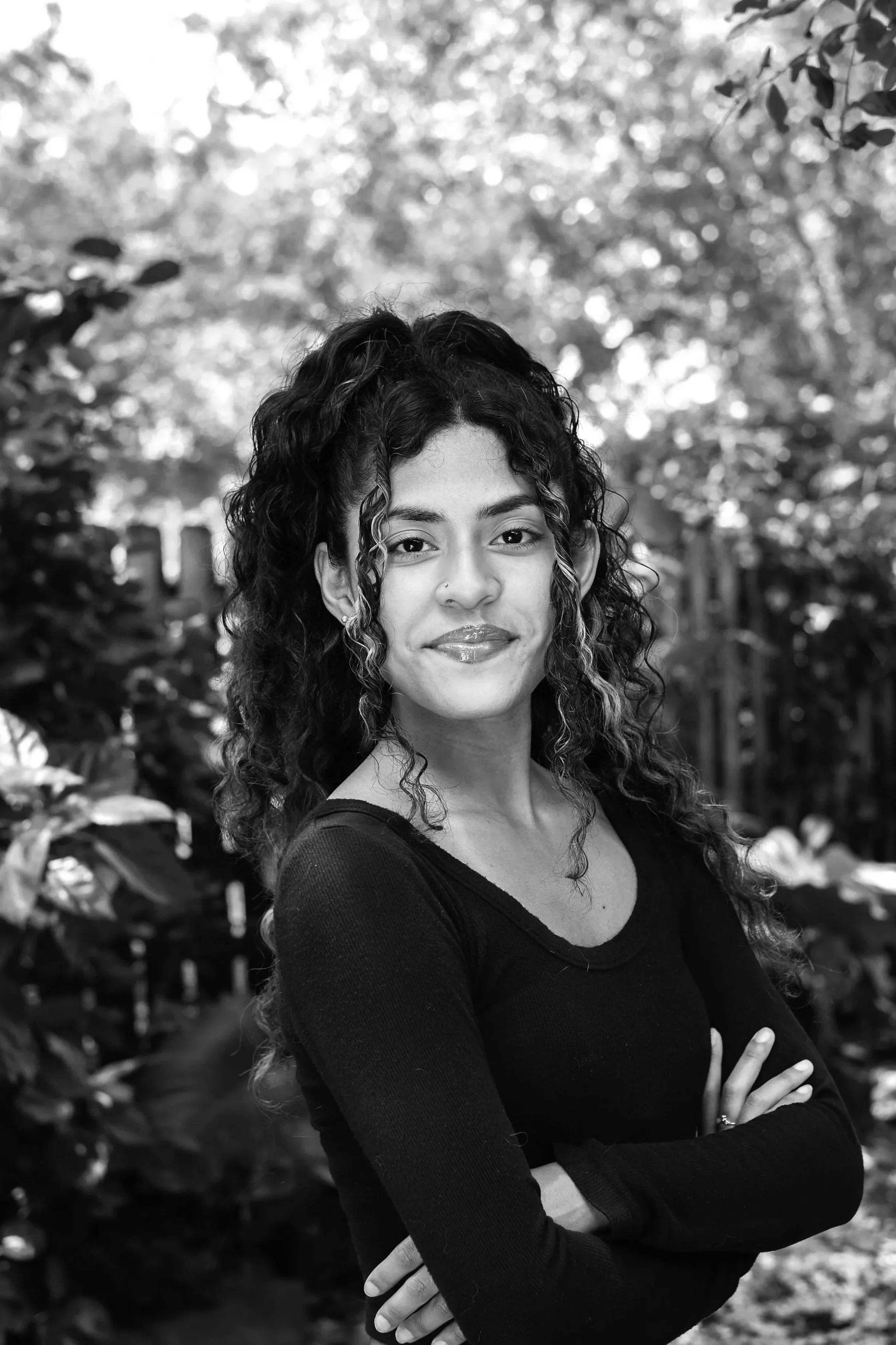 A black-and-white portrait of a young woman with curly hair, arms crossed, smiling slightly, outdoors with trees and foliage in the background.