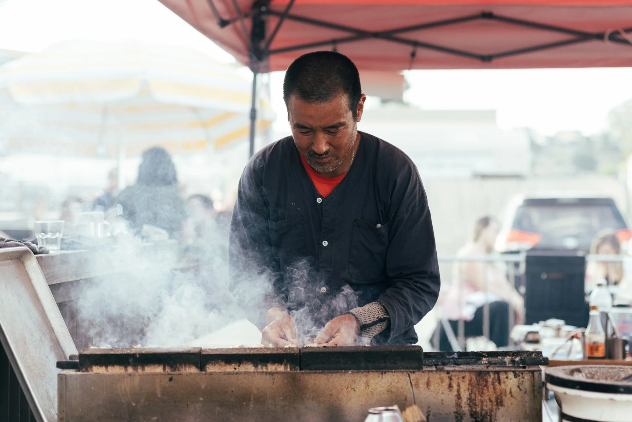 A man cooking on a grill at an outdoor food stall, surrounded by smoke, with a red canopy overhead and other people in the background.