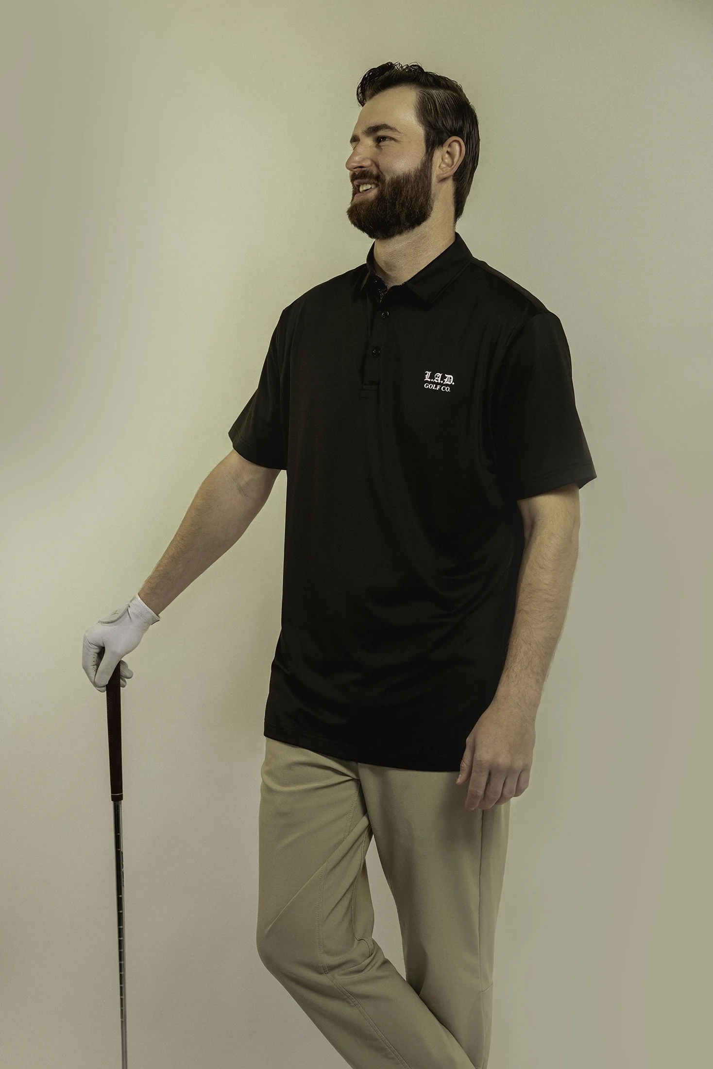A man with a beard wearing a black polo shirt with 'LAD GOLF CO.' logo, beige pants, and a white golf glove, holding a golf club, standing against a plain off-white background.