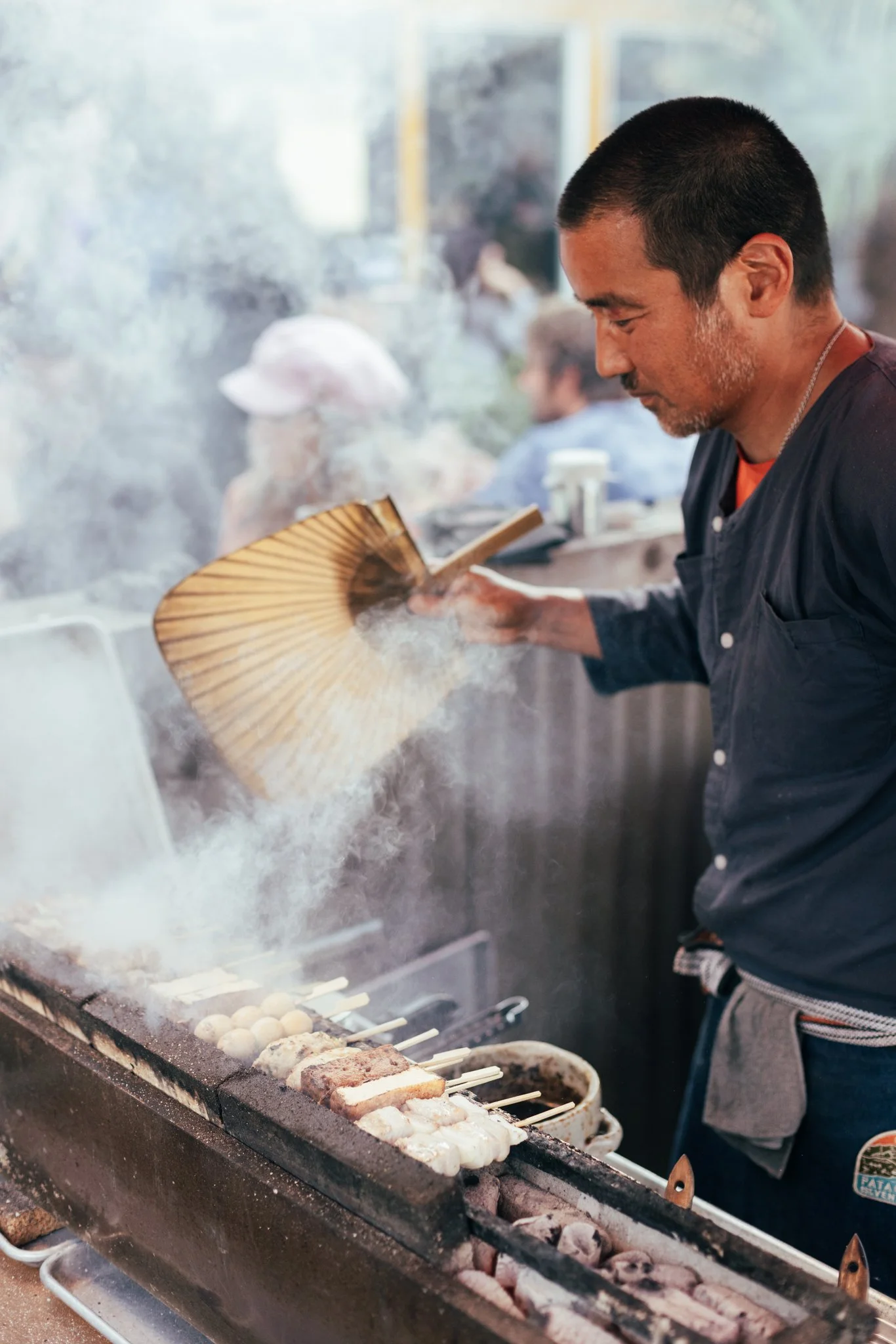 A man grilling skewers of meat and vegetables over an open flame at a street food stall, with steam and smoke rising.