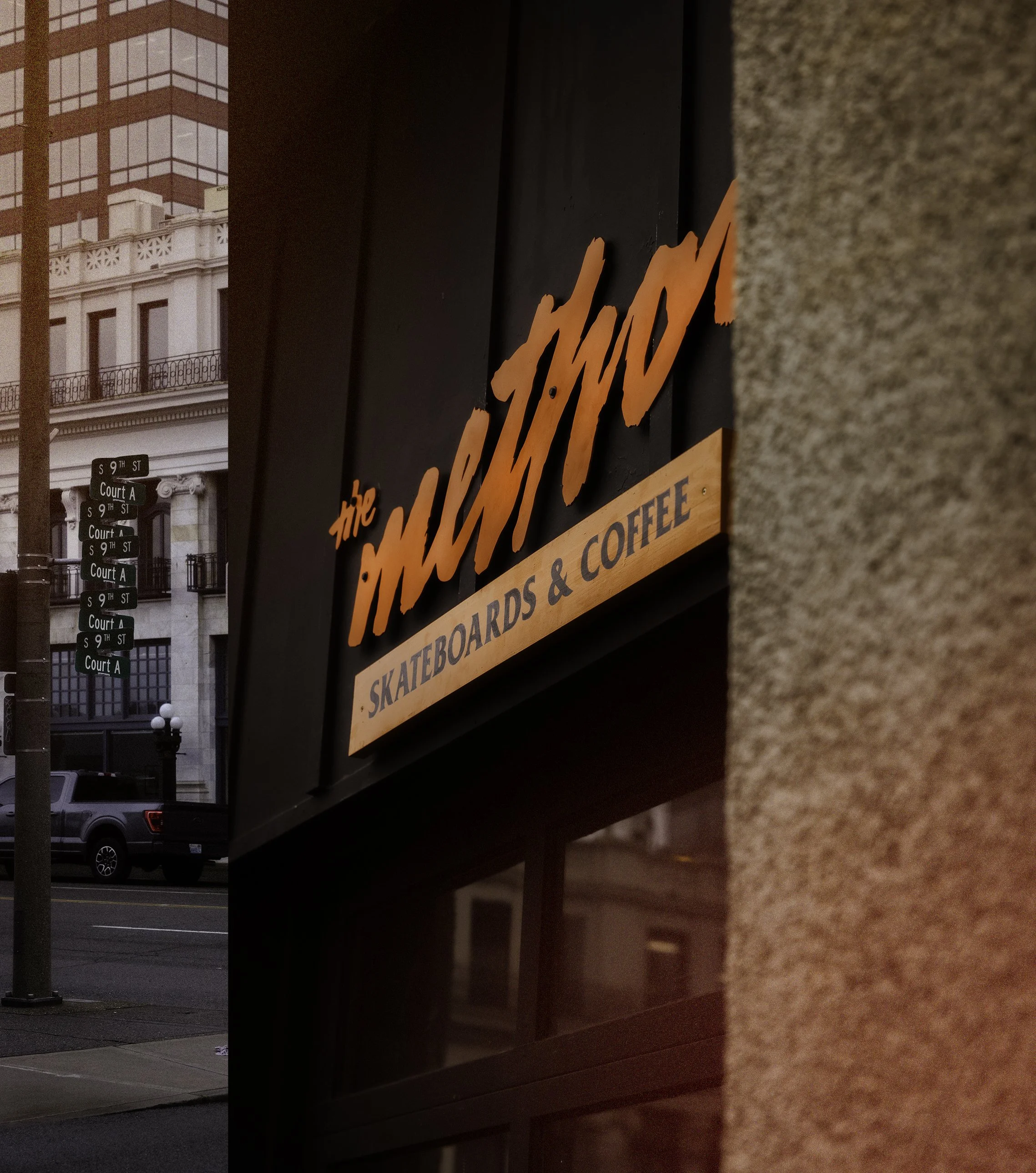 A neon sign for 'the MAKERSPACE' skateboards and coffee, mounted on a black wall outside a building, with part of a stone wall in the foreground and city street with cars and buildings in the background.