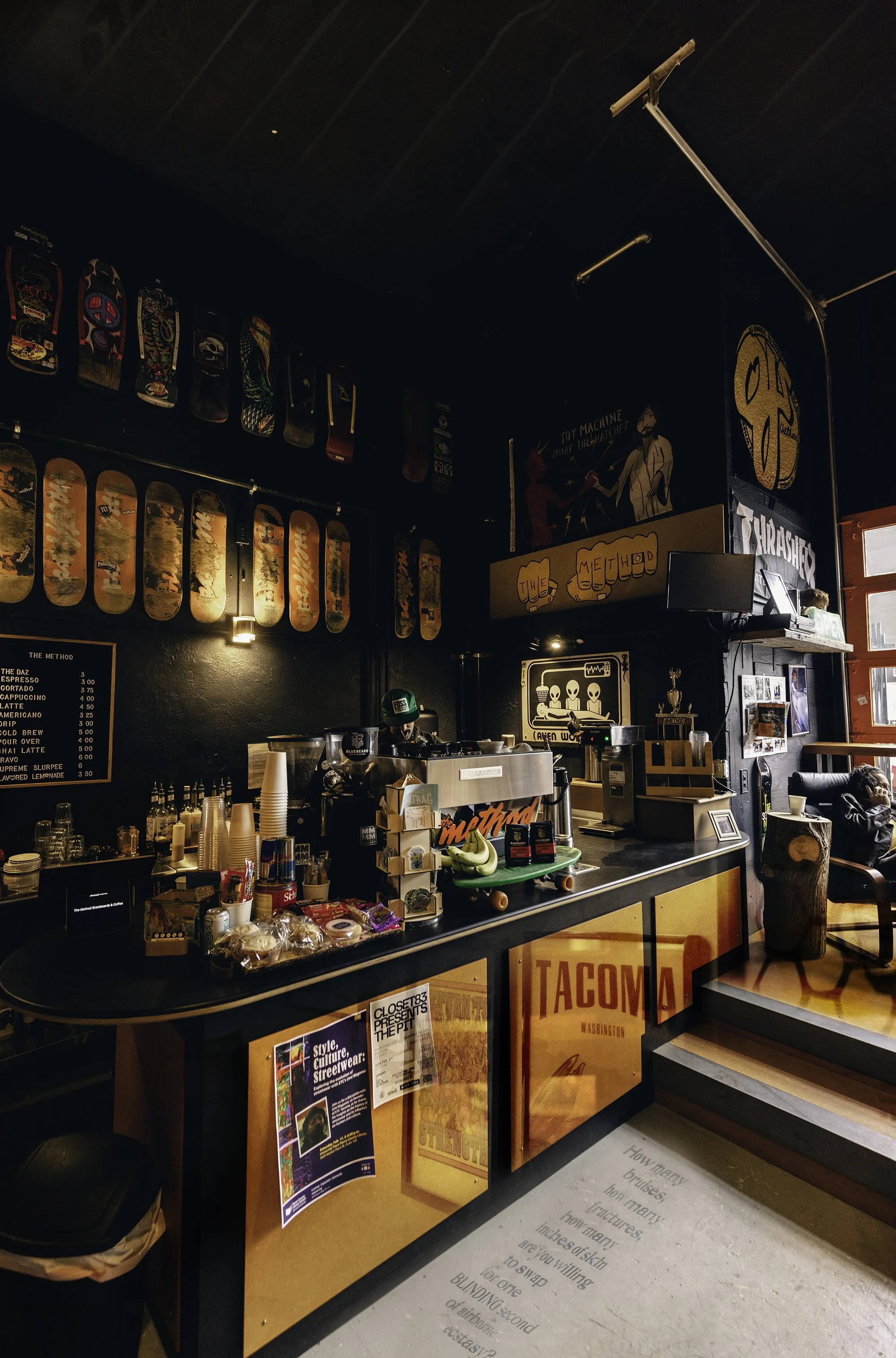 Inside a coffee shop with skateboard art on the walls, a colorful counter with various snacks, cups, and a coffee machine, a person wearing a green cap preparing a drink, and a cozy seating area to the right.