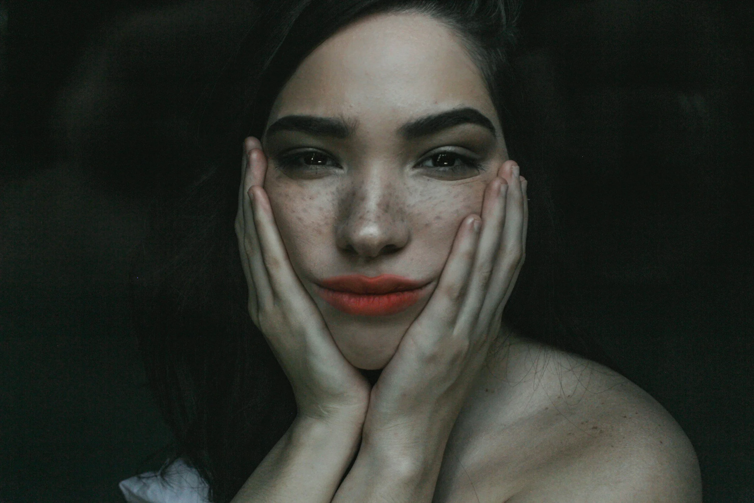 A woman with dark hair, freckles, and full lips resting her chin on her hands against a dark background.
