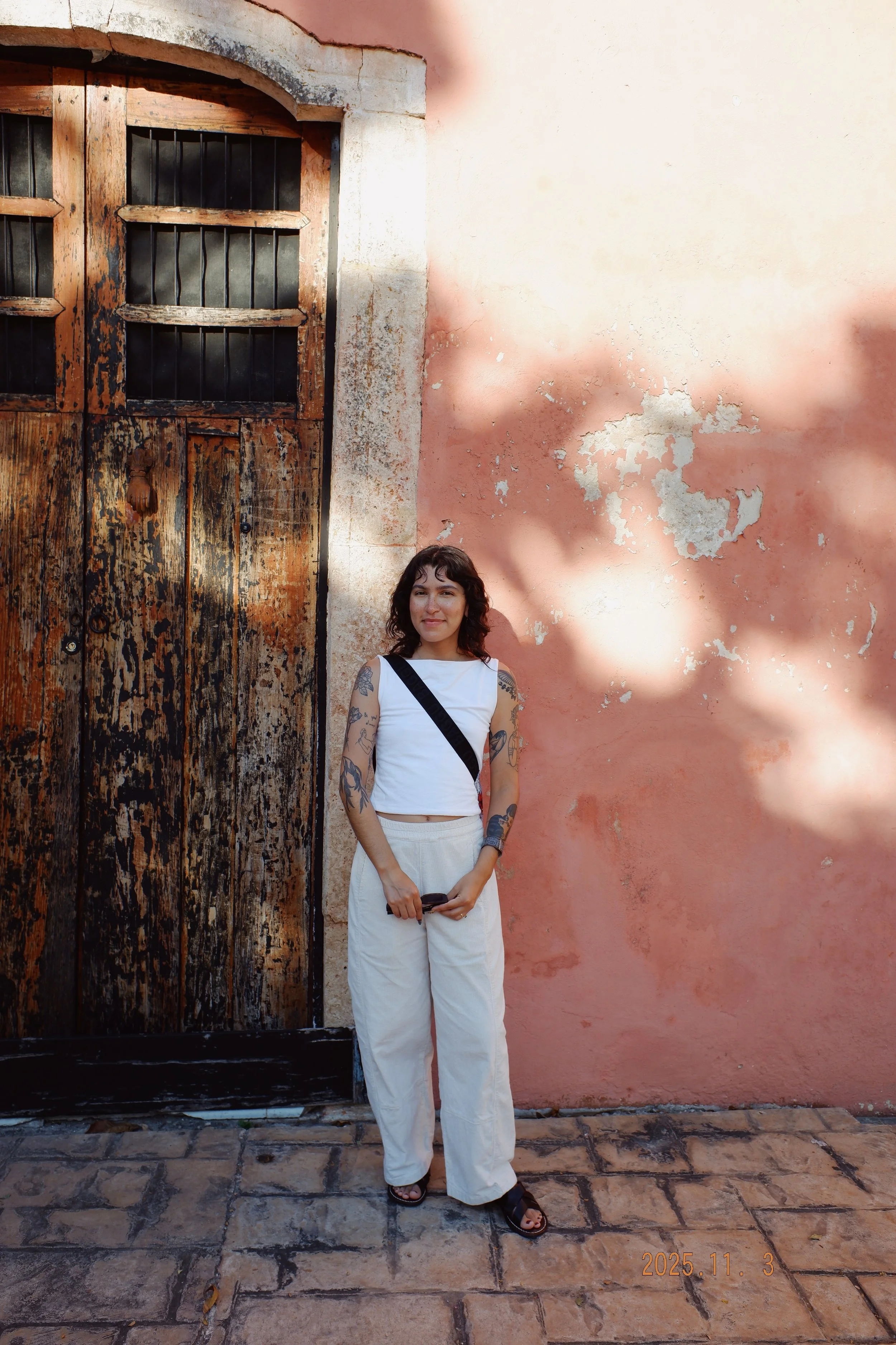 A person with tattoos standing in front of an old wooden door and a textured pink wall with peeling paint, casting shadows on the wall in Mexico.