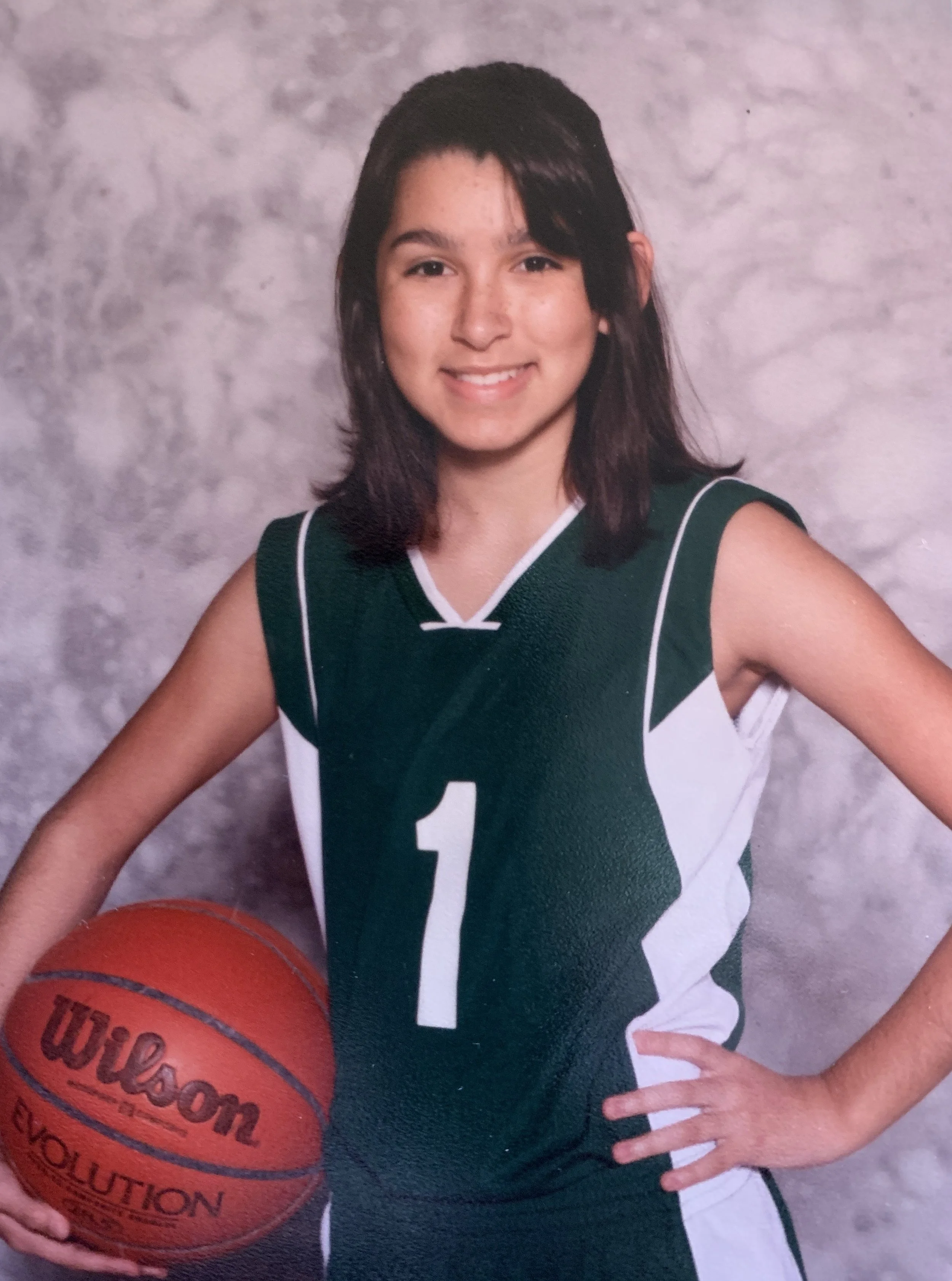 Young girl in a green and white basketball uniform holding an orange Wilson basketball, smiling, against a gray textured background.