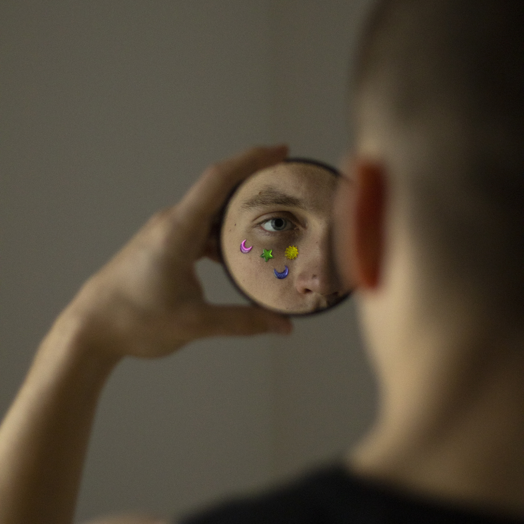 A person holding a small round mirror, showing their face reflected with colorful star, moon, and sun stickers covering their face, while their eye looks into the mirror.