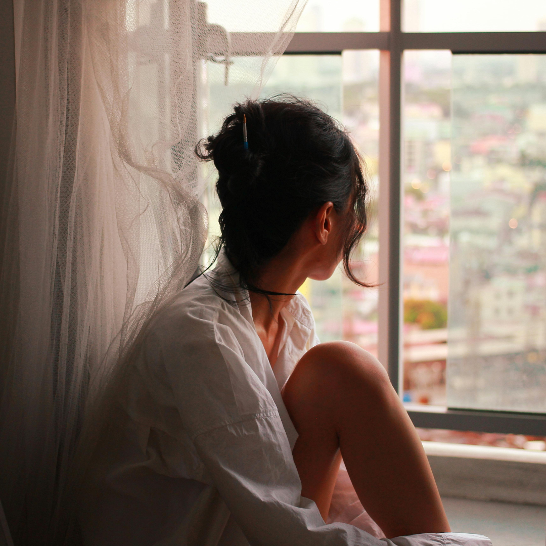 A woman sitting by a window, looking outside, with her knee drawn up. She wears a white shirt and has dark curly hair tied up, with a pen tucked in her hair. She looks contemplative and is near a curtain, with a cityscape visible through the window.