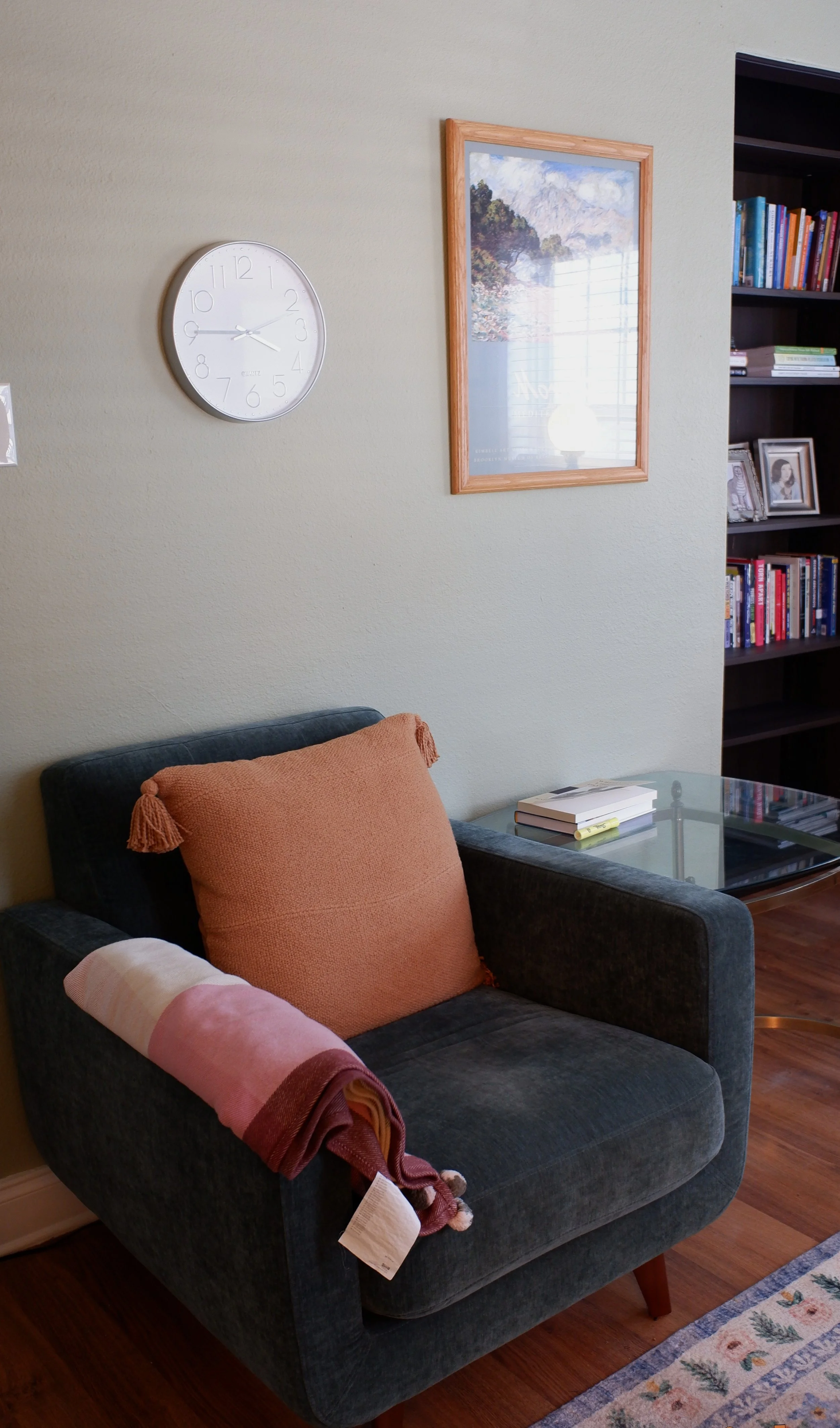 Living room with a gray armchair, orange cushion, pink and white throw blanket, glass-top side table with books, white wall clock, framed landscape picture, and a bookshelf filled with books.