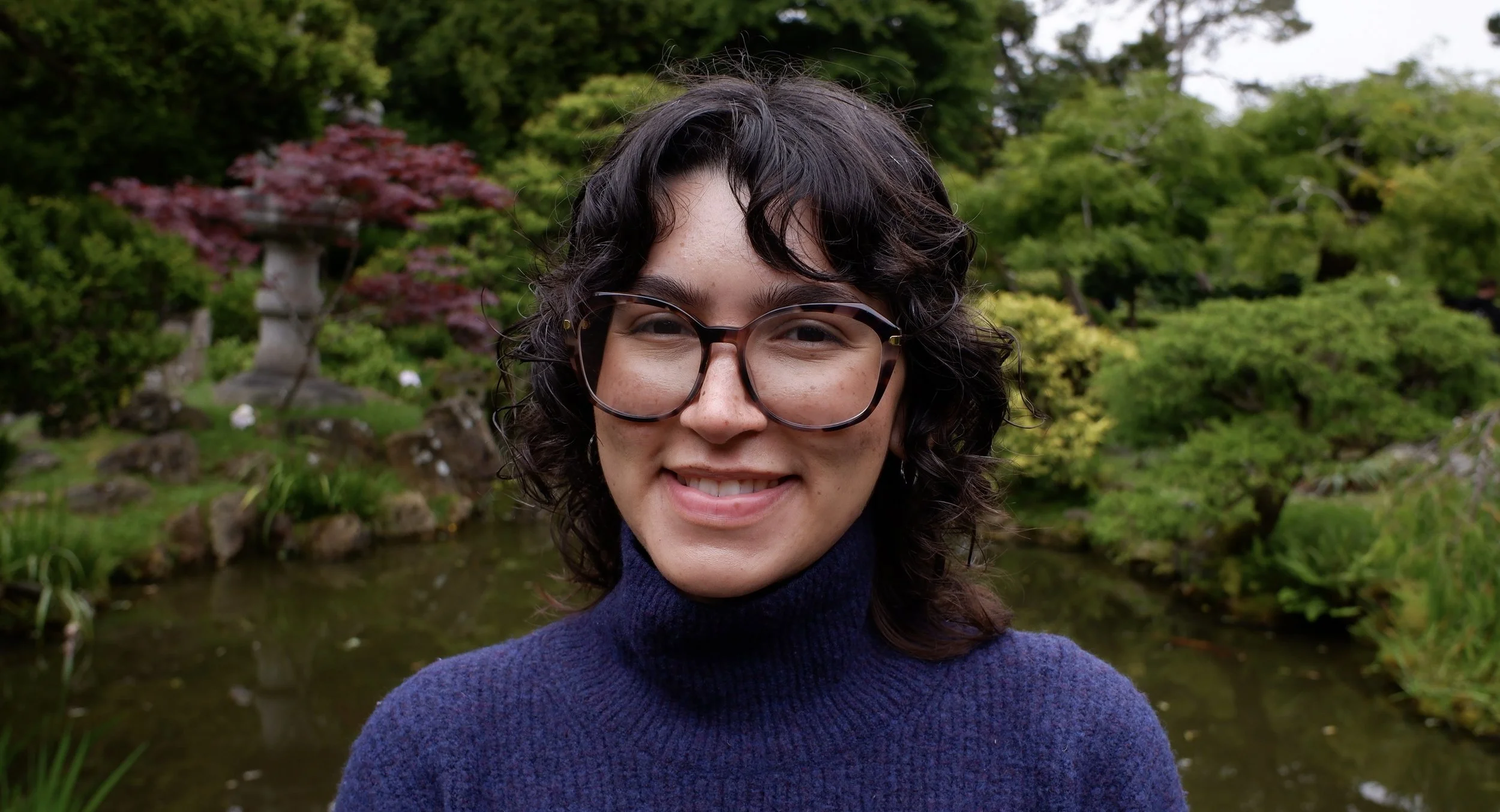 Close-up of a smiling person wearing glasses and a navy blue turtleneck sweater, standing in front of a lush garden with trees, rocks, and a pond.
