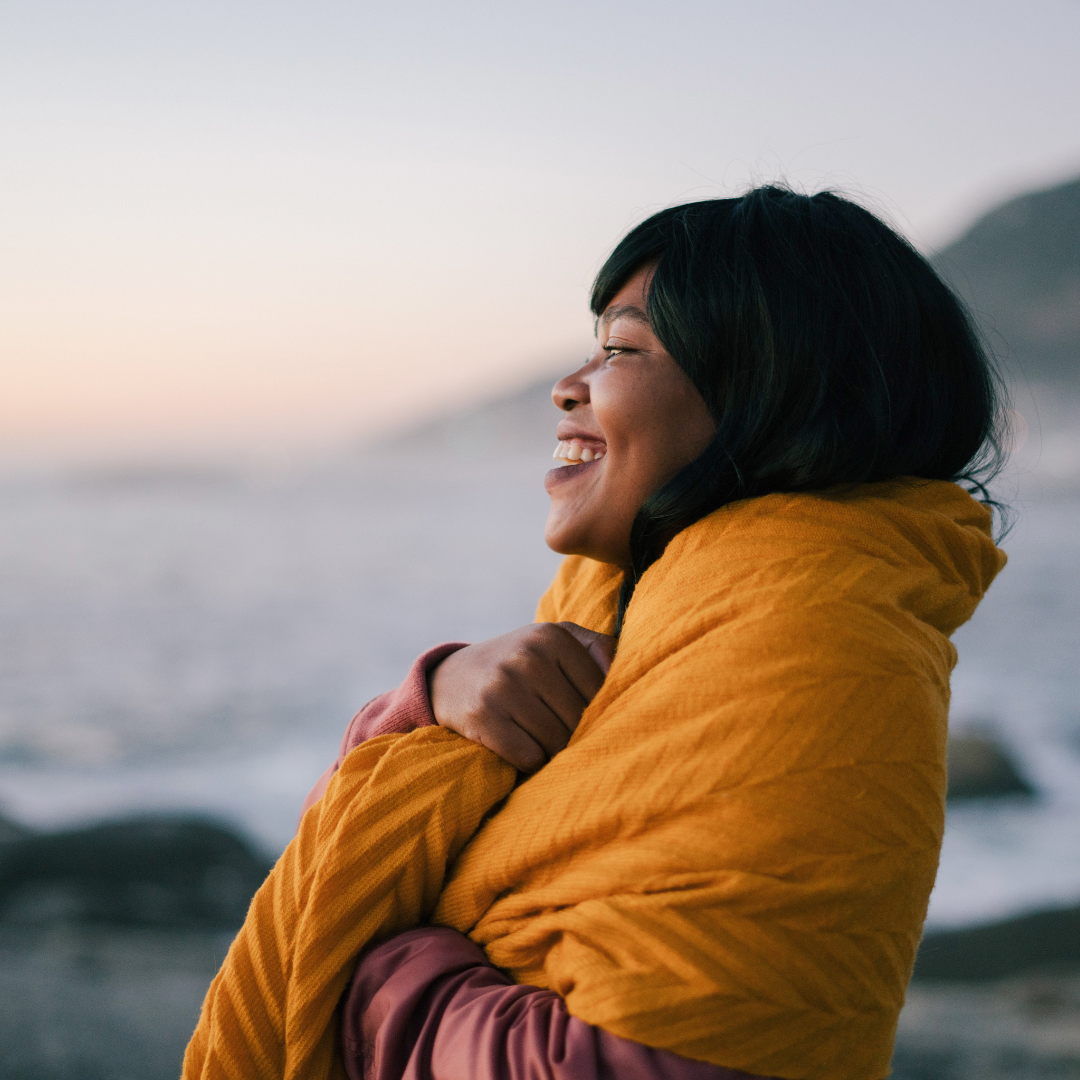 A smiling woman with dark hair wrapped in a yellow blanket stands outdoors near the ocean during sunset.