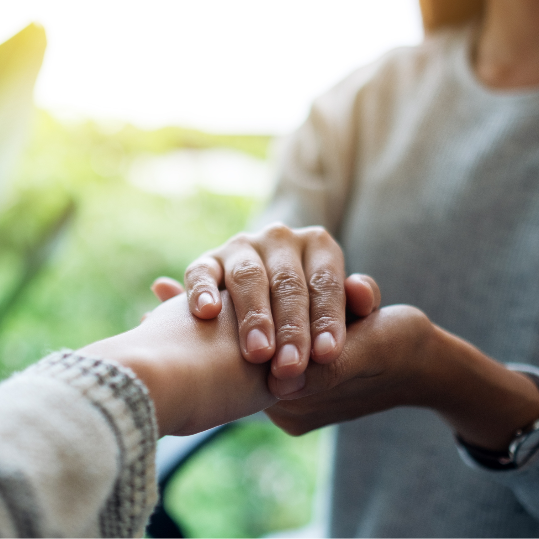 Two people holding hands, one person gently gripping the other's wrist in a caring gesture outdoors with sunlight and blurred greenery in the background.