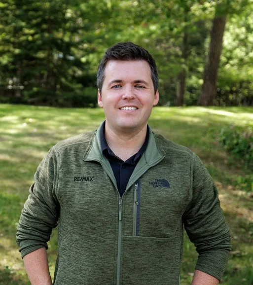 A man standing outdoors in a green park, wearing a green North Face jacket and smiling at the camera.