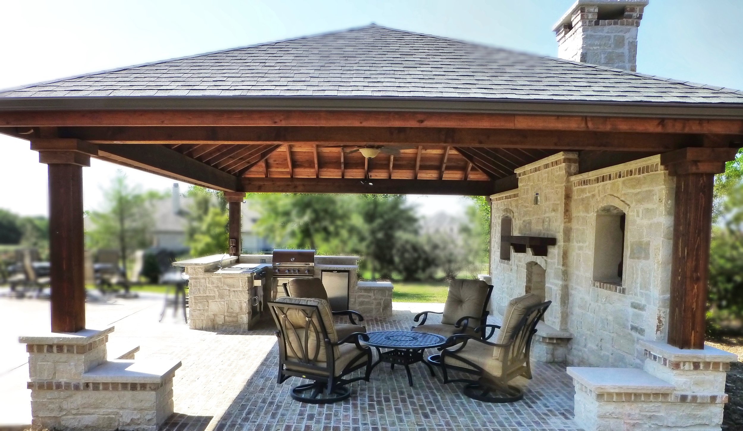 Outdoor covered patio with stone brick flooring, wooden beams, outdoor chairs around a small round table, built-in brick grill, and a stone fireplace, with trees in the background.