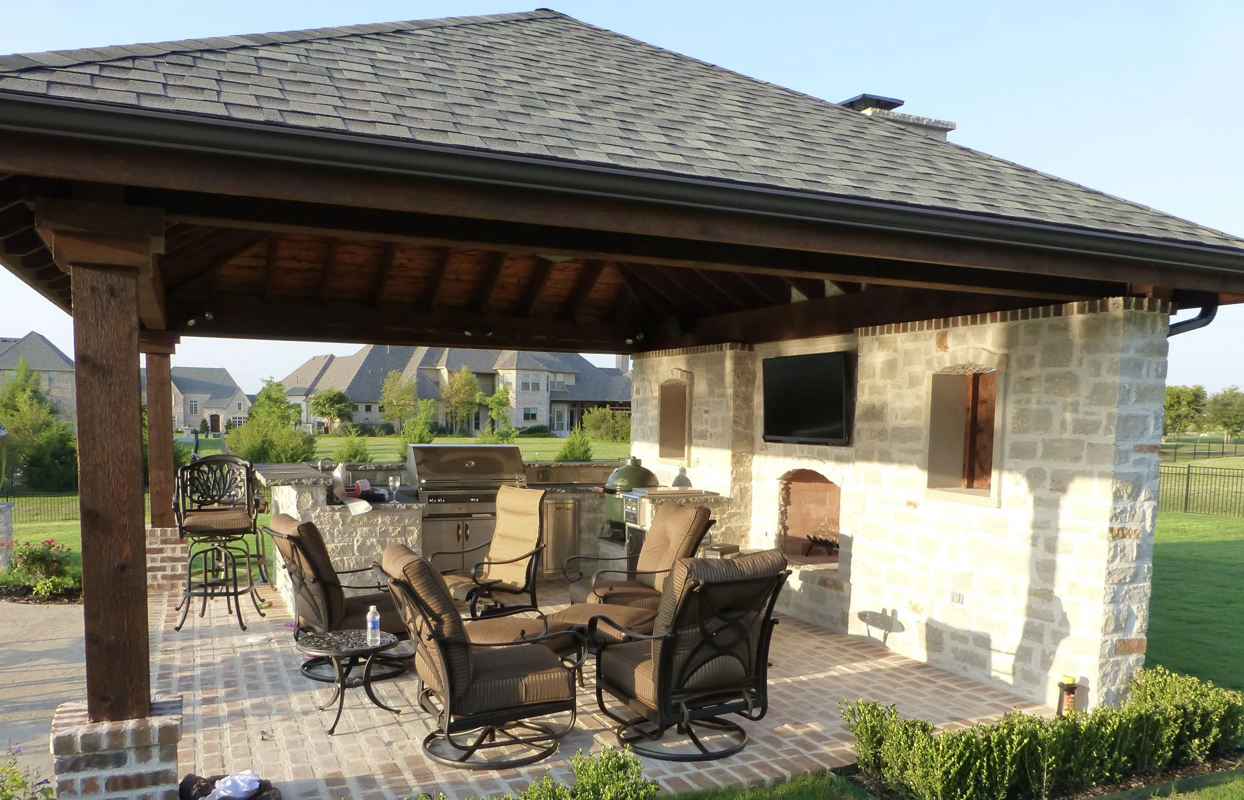 Outdoor covered patio with a brick floor, a stone fireplace, a wall-mounted TV, a barbecue grill, a table with six cushioned chairs, and wooden posts supporting the roof. In the background, there are houses and a lawn.