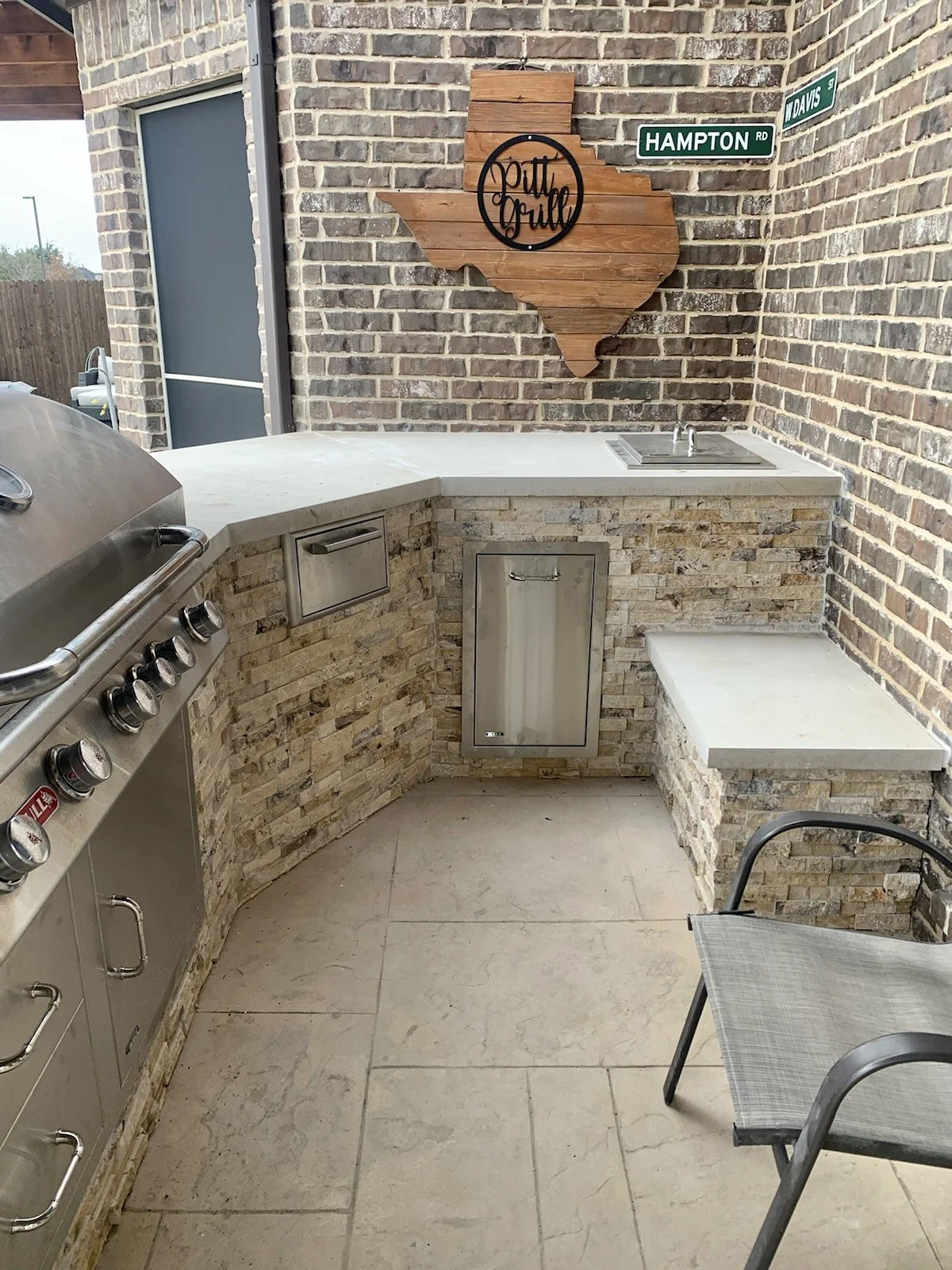 Outdoor kitchen area with brick walls, featuring a stainless steel grill, sink, and a wooden sign shaped like Texas with "Pit Grill" written on it. There are metal signs reading "Hampton Rd" and "Davis St" on the brick wall.