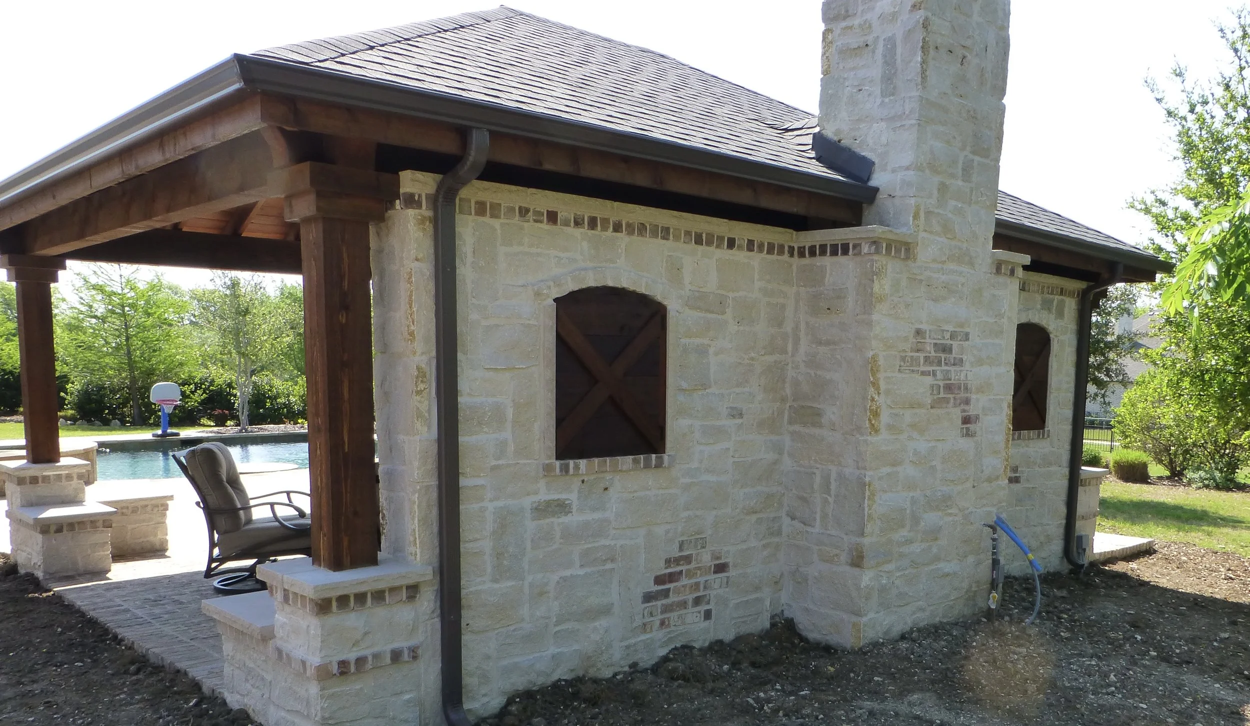 Backyard patio with stone and brick outdoor structure, wooden beams, patio furniture, and a swimming pool in the background.