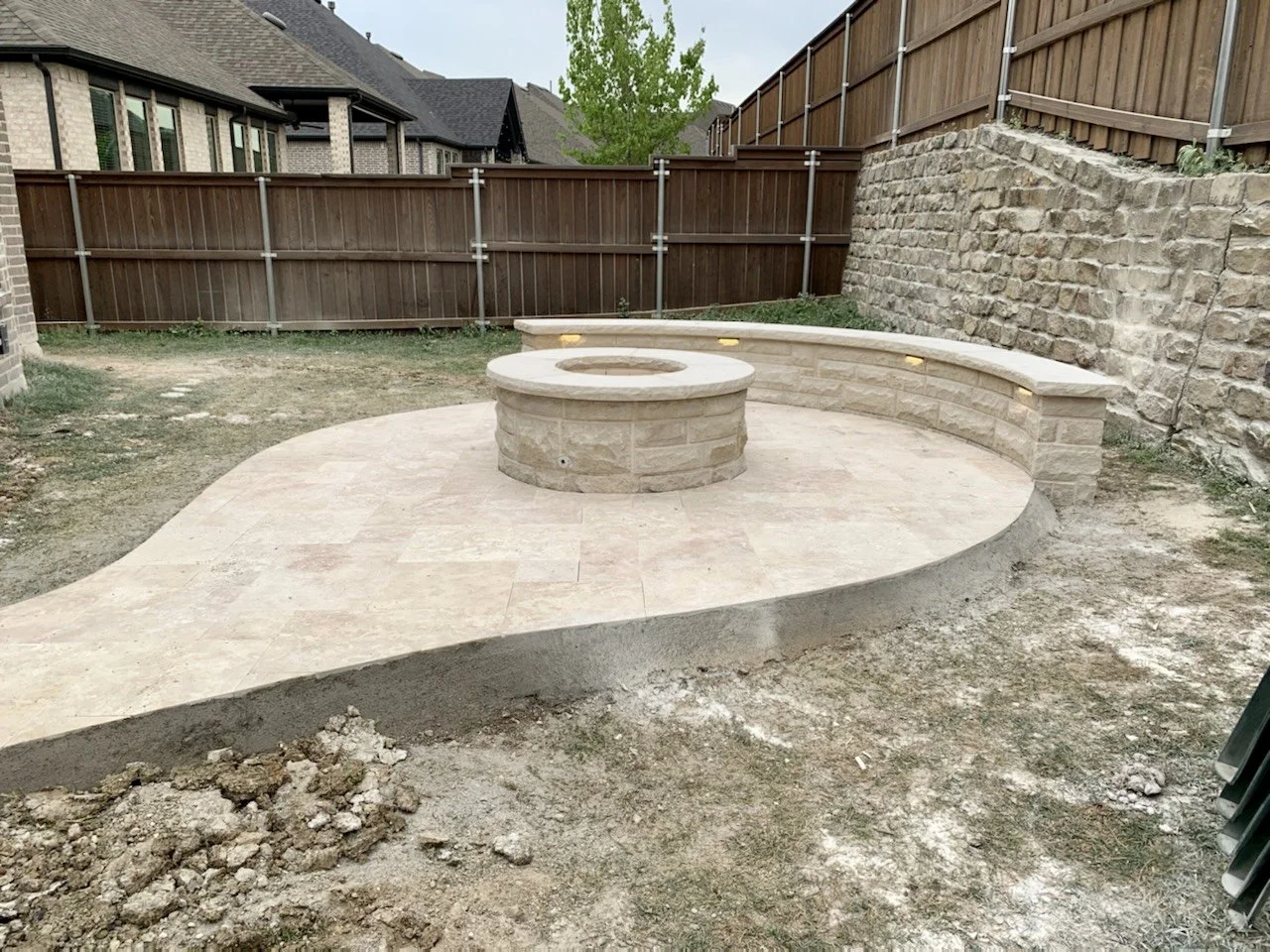 Backyard with stone patio featuring a built-in bench and circular fire pit, surrounded by wooden fencing and stone retaining wall.