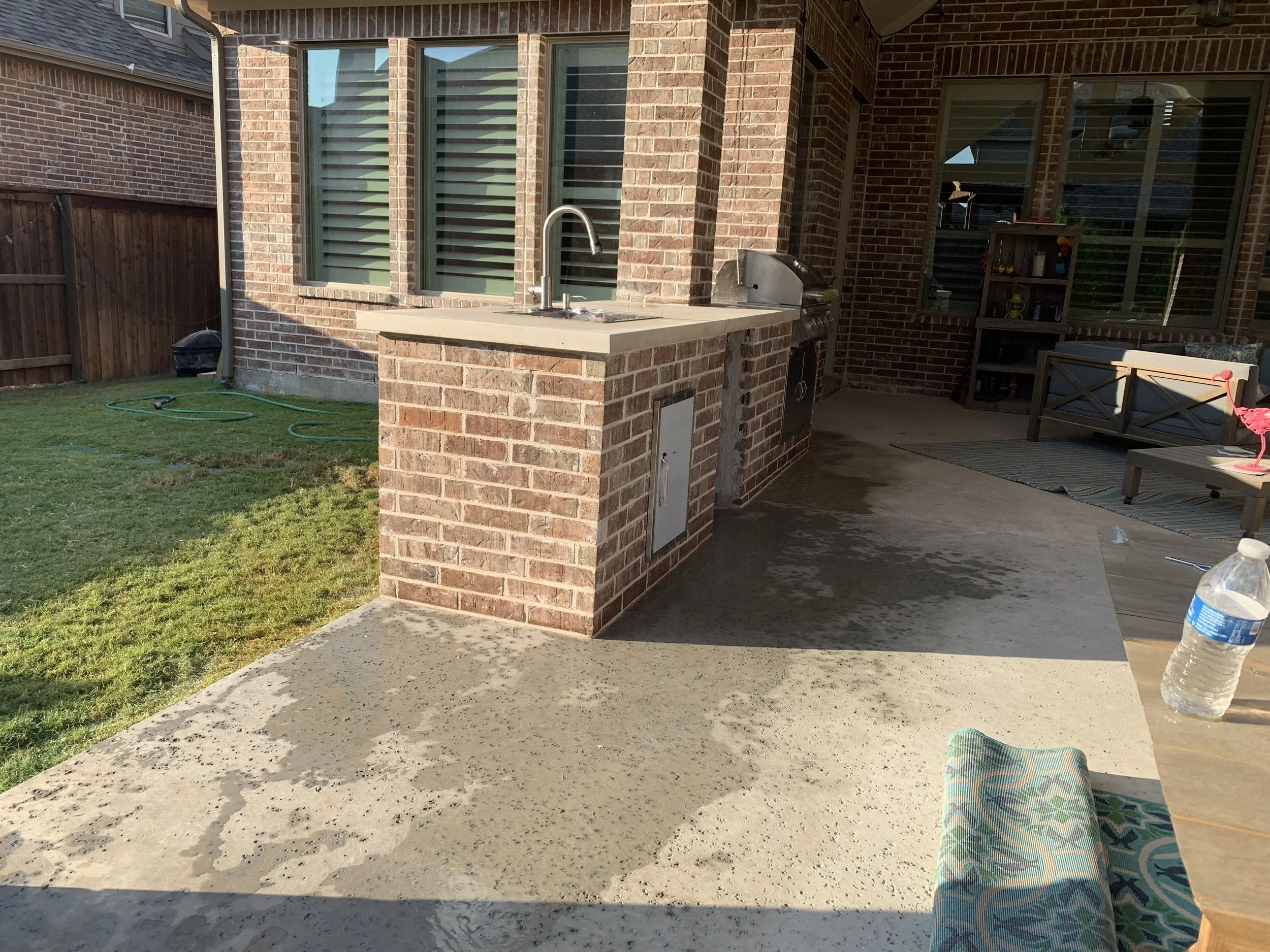 Backyard patio with brick outdoor kitchen station featuring a small sink, grill, and a closed cabinet. There is a wooden outdoor bench with pink flamingo decoration on it, a water bottle on the table, and a rolled-up outdoor rug. The patio is adjacent to a grassy yard and is enclosed by a wooden fence.