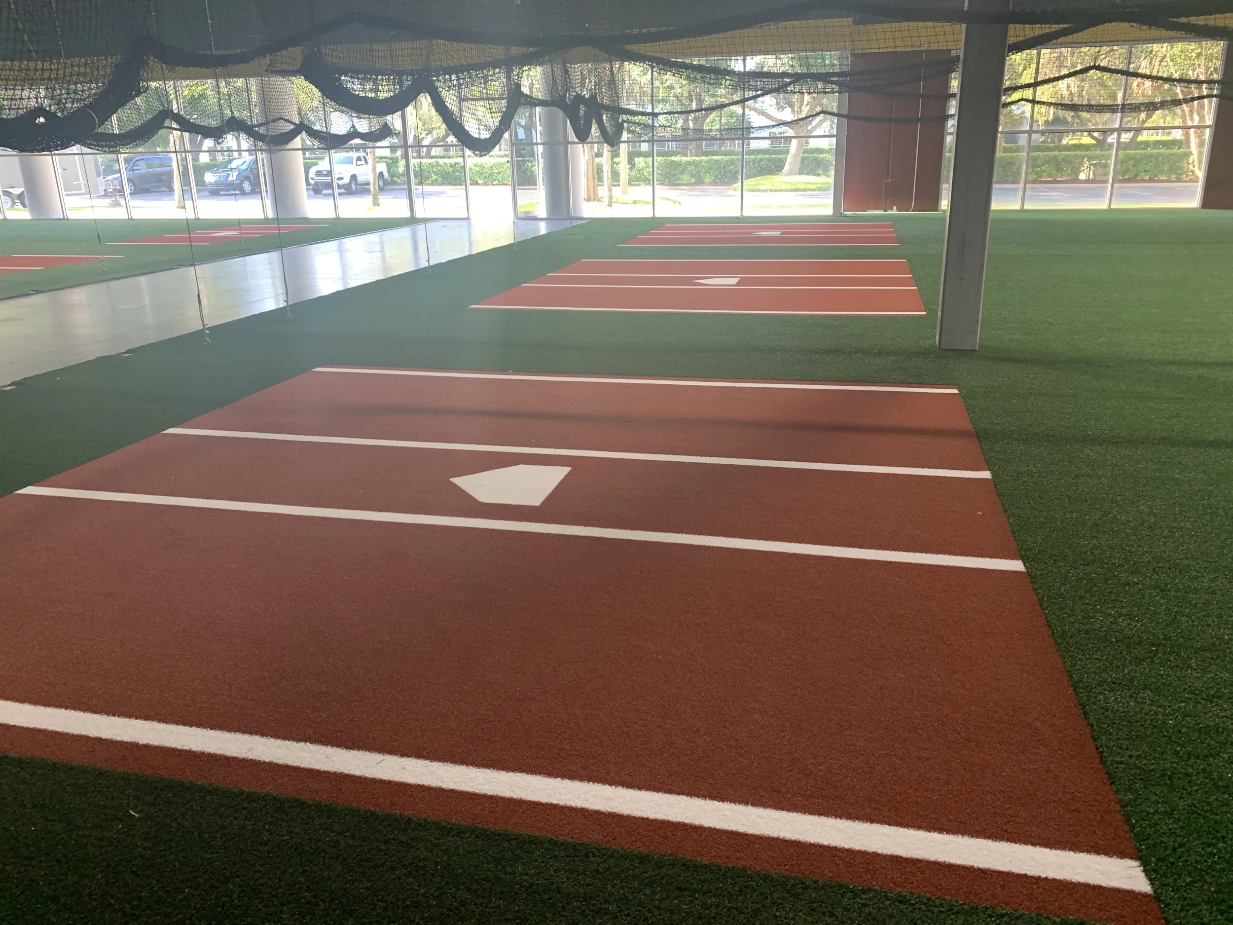 Indoor batting cages with artificial turf and pitching lanes, featuring white home plate markers, surrounded by netted walls.